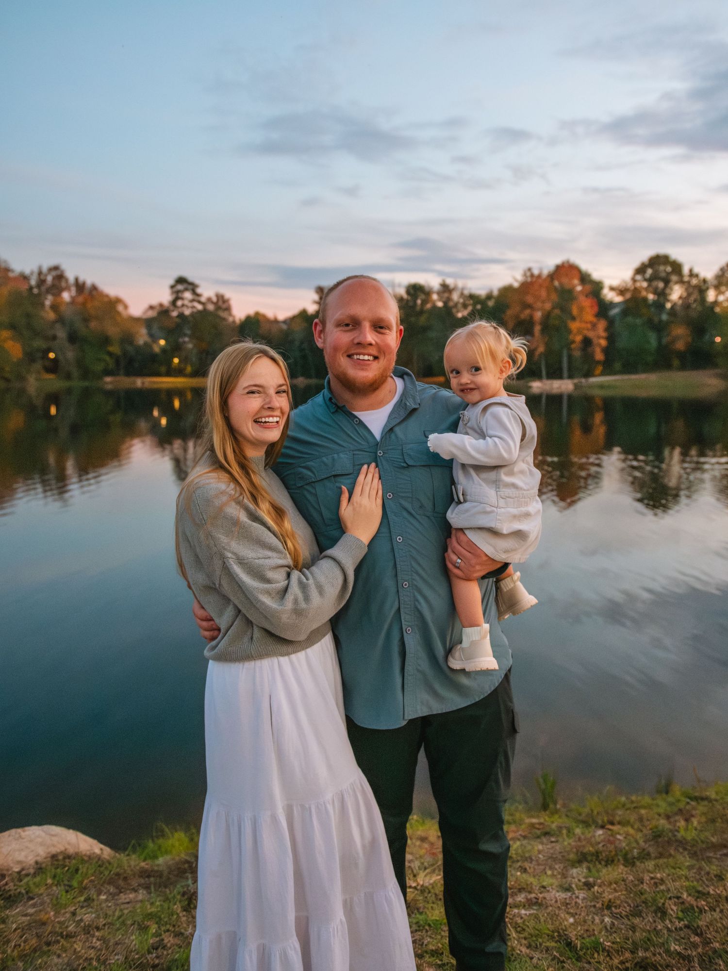 Family of three smiling together by a lake during a golden hour outdoor session. At the Lexington Old Mill Pond