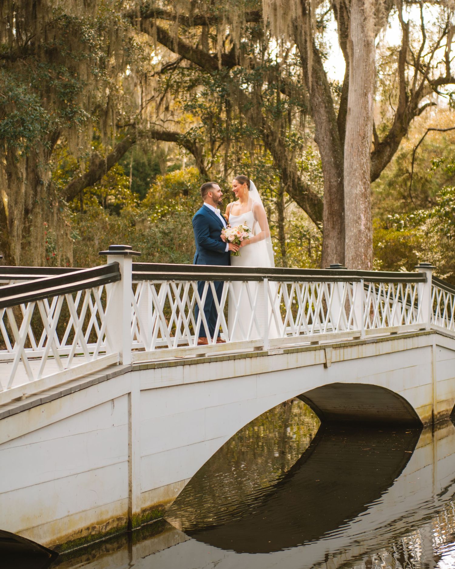 Bride and groom standing on a white bridge surrounded by trees during a romantic wedding portrait. Magnolia Plantation and Gardens