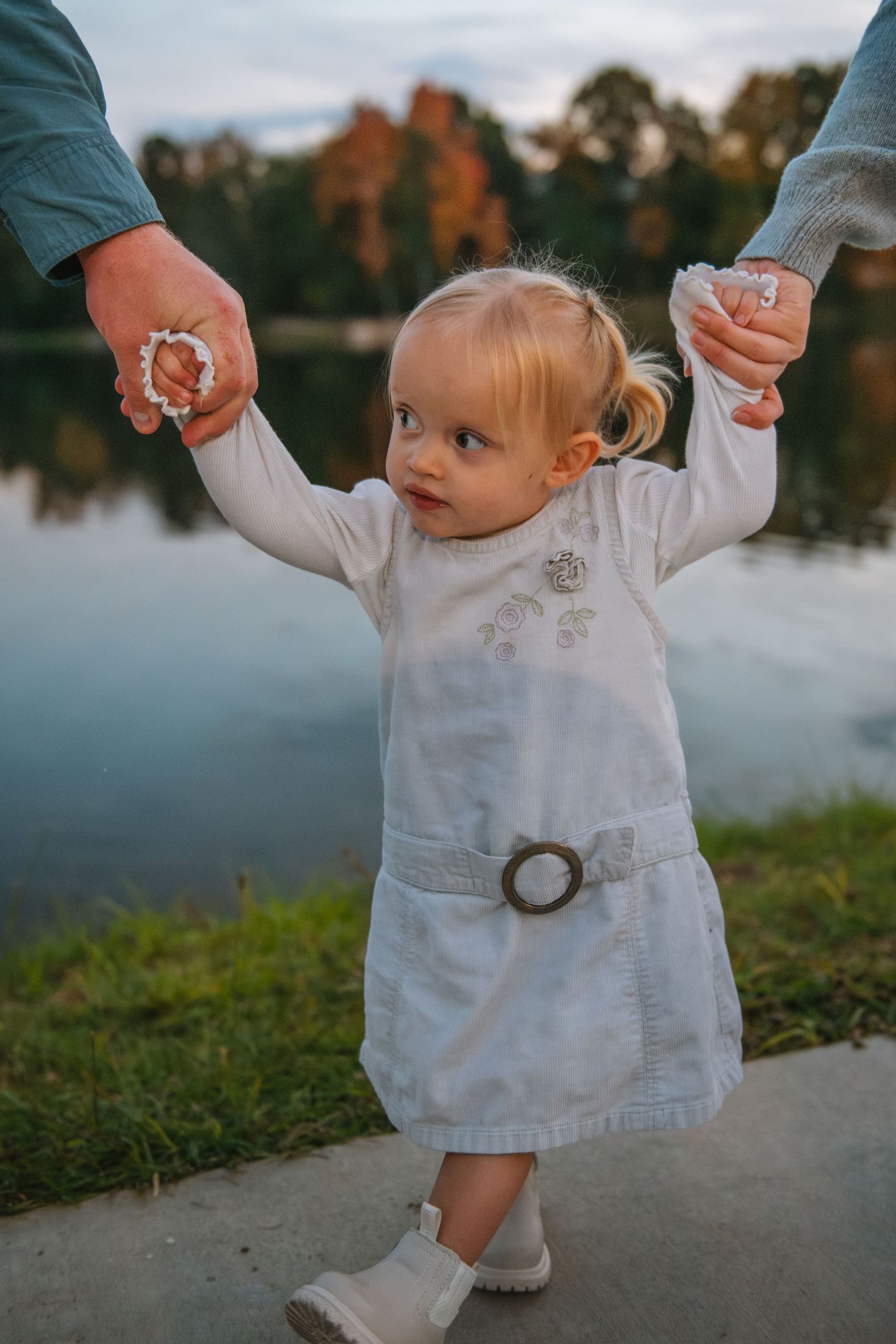 Baby girl taking first steps while holding parents hands during a lakeside family session in South Carolina. At the Lexington Old Mill Pond