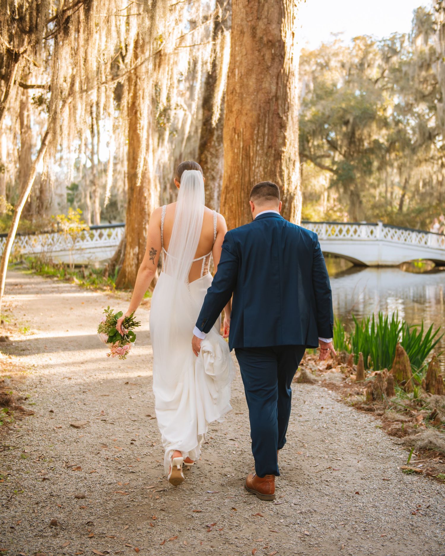 Bride and groom walking hand in hand along a scenic path under Spanish moss in a South Carolina wedding setting.