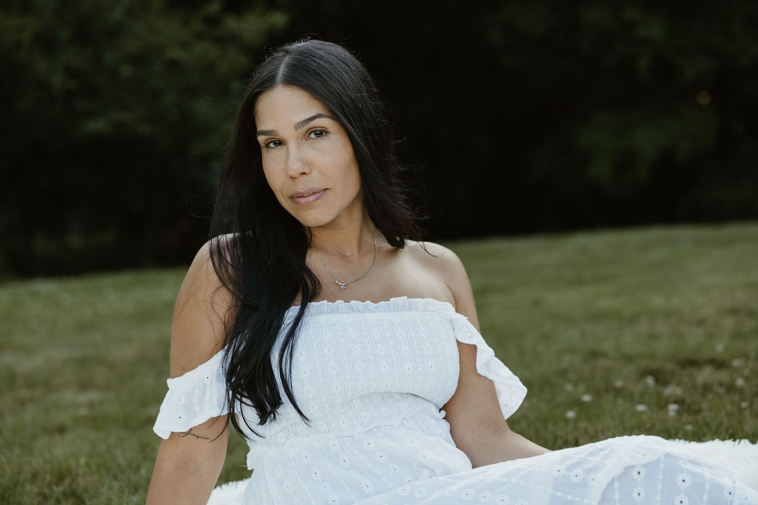 Moody outdoor portrait of woman in white off-shoulder eyelet dress seated on grass with dark tree backdrop in Woodbridge CT