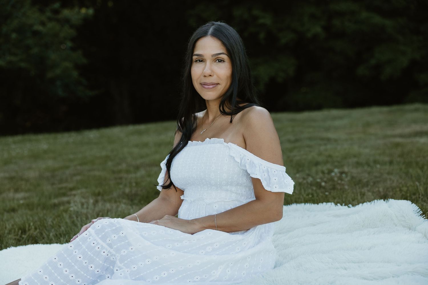 Editorial portrait of woman in white off-shoulder dress seated outdoors on green lawn at golden hour in Woodbridge CT