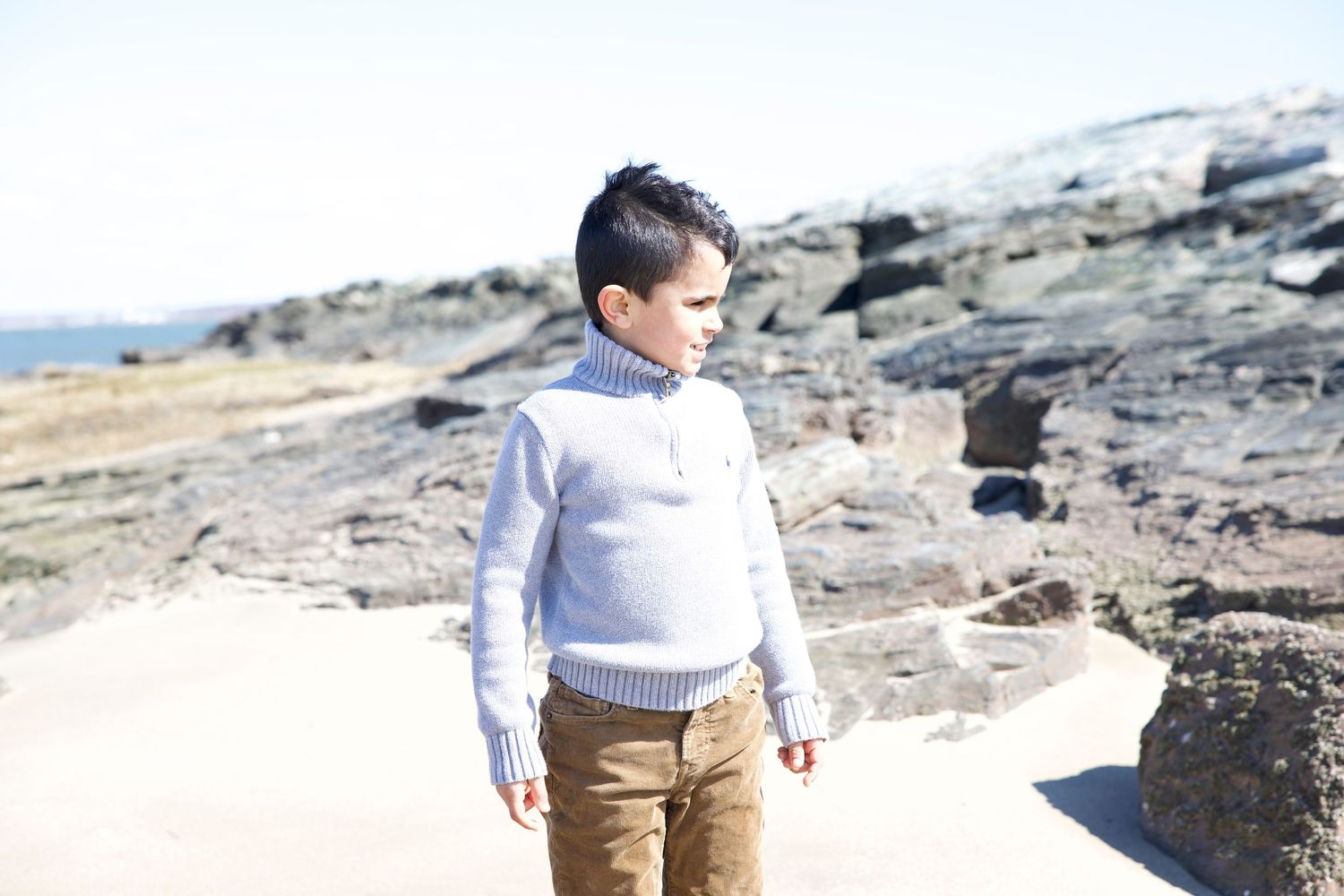 Young boy in pale gray sweater standing on rocky beach at Sea Bluff Beach, color portrait by Noemi Blumenthal Photography