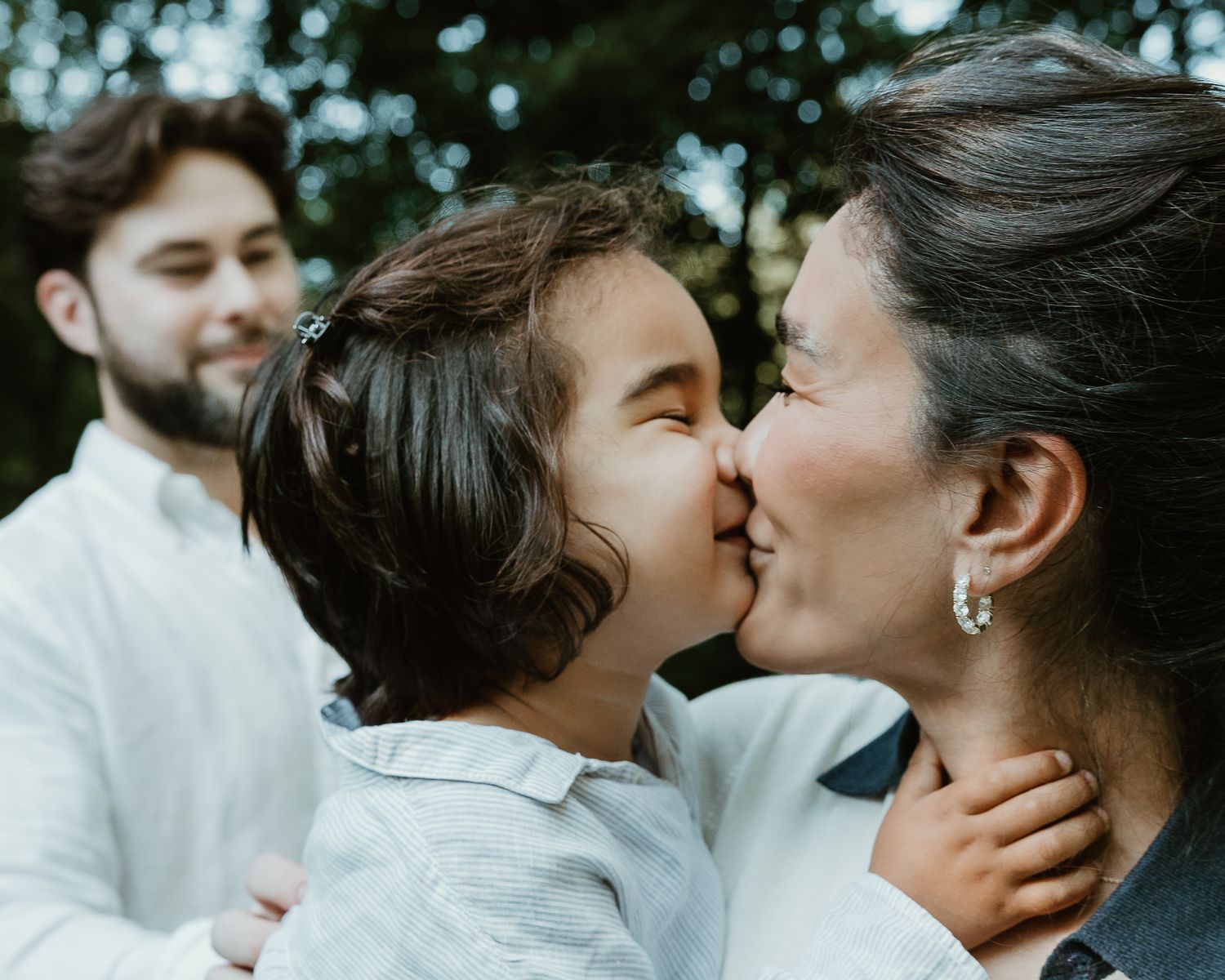 Mother kissing young child's cheek while father looks on, outdoor family portrait in Woodbridge, CT