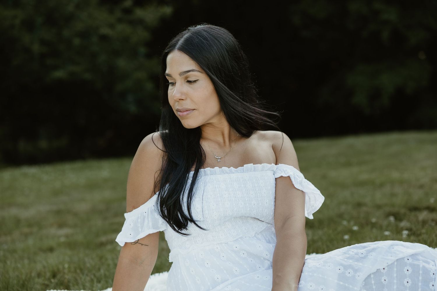 Candid portrait of woman in white off-shoulder eyelet dress looking to the side while seated on grass in Woodbridge CT