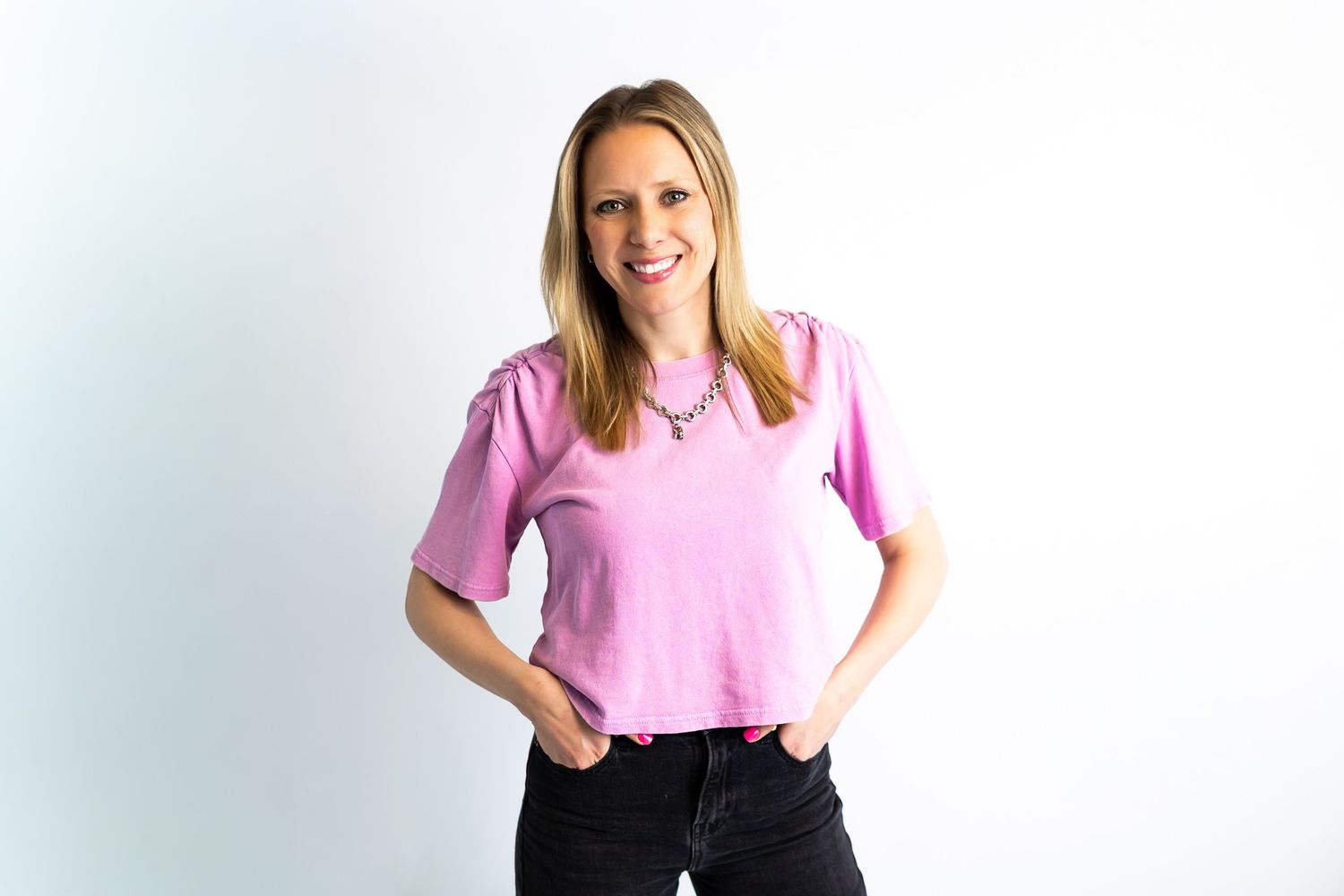 A person in a pink t-shirt and black pants stands smiling against a light background.