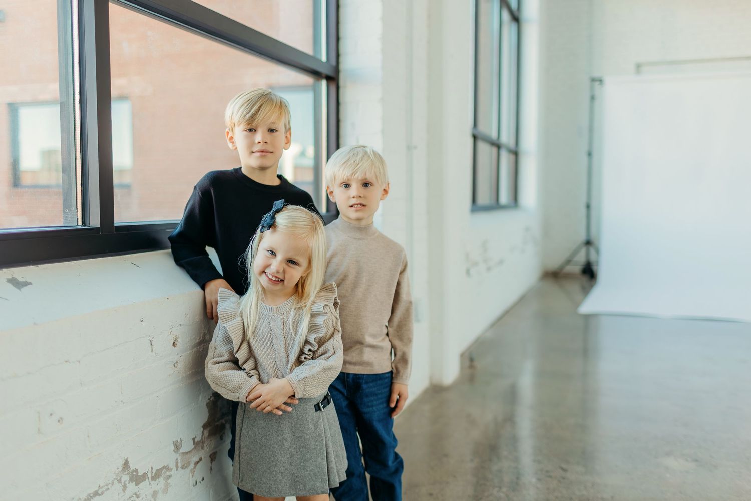 Children in neutral colored sweaters standing by bright windows in modern space.