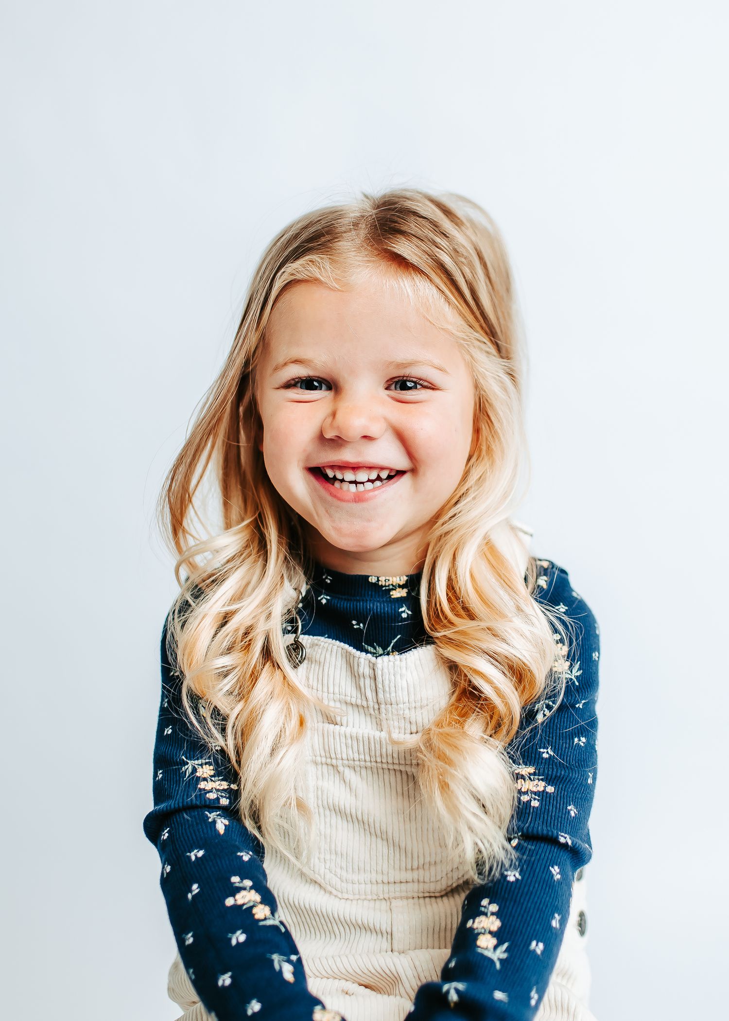 Child wearing a navy blue floral top and cream colored overalls smiles brightly against a white background.