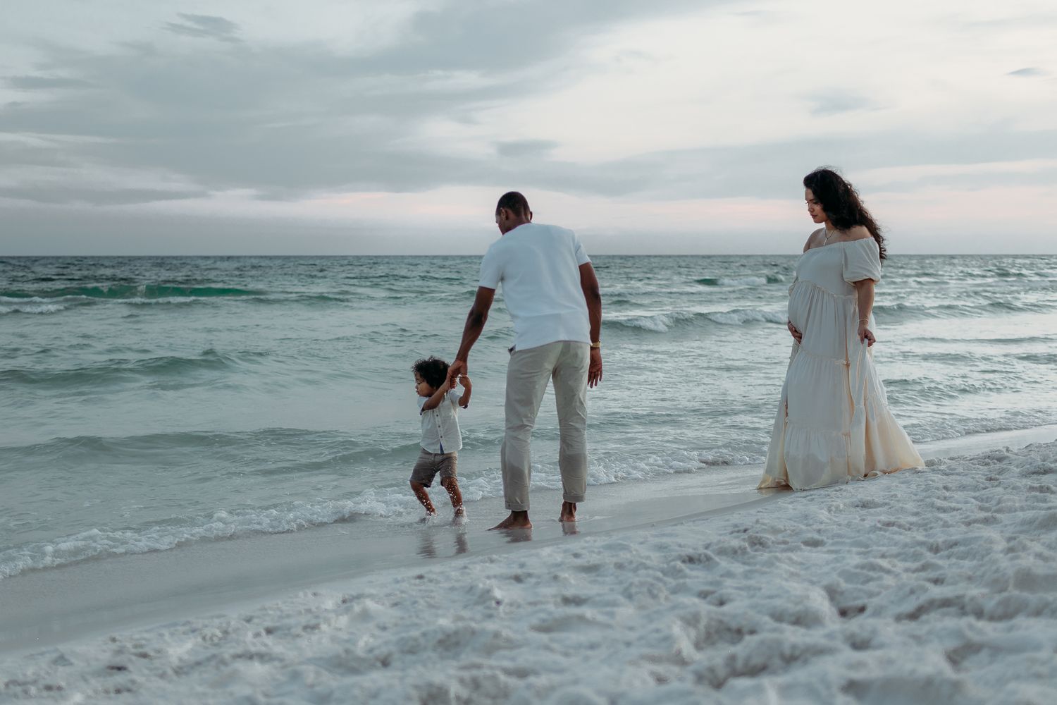 Family members walking and playing in the ocean waves at sunset along a white sandy beach.