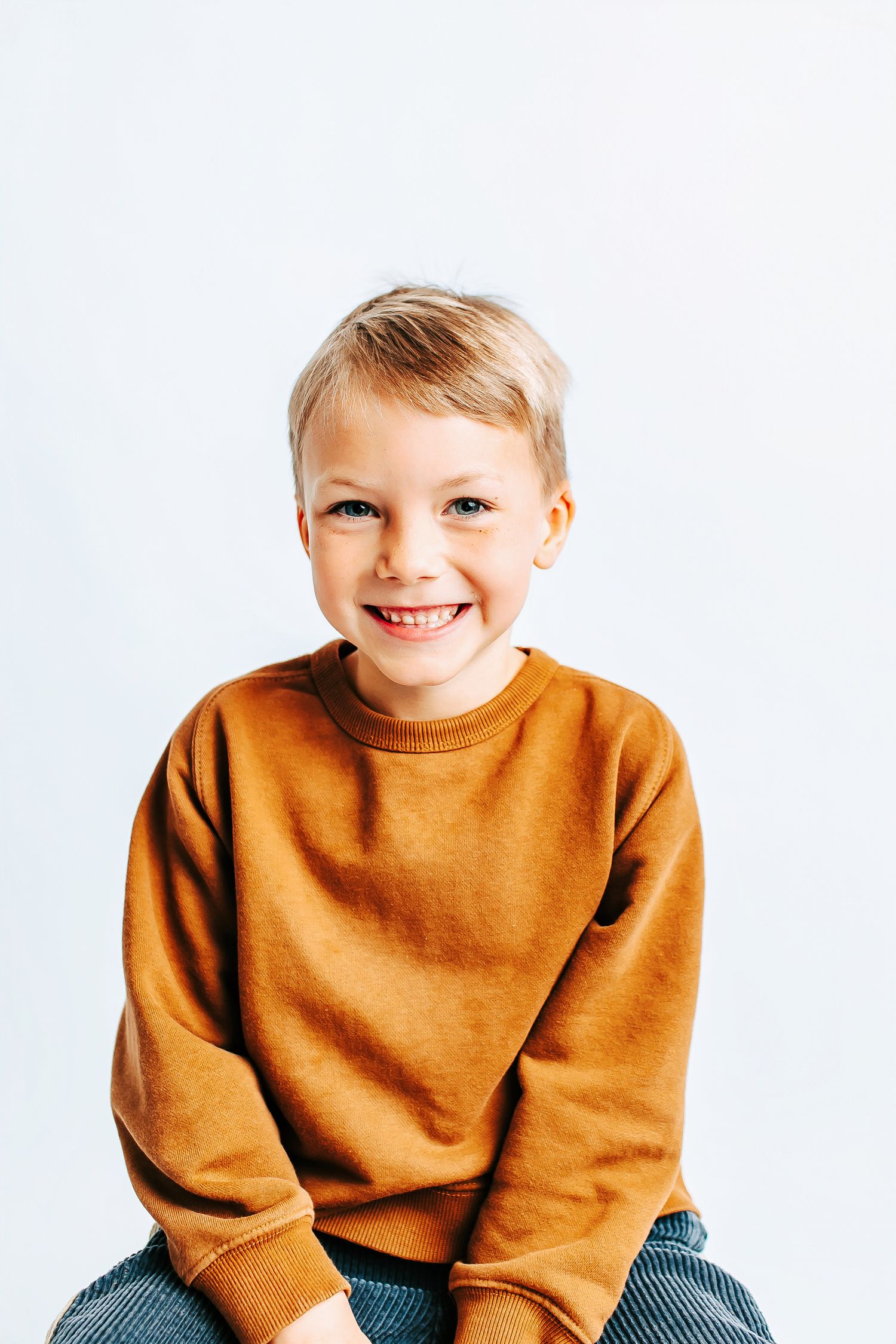 Young child wearing a cozy brown sweater sits and smiles against a white background.