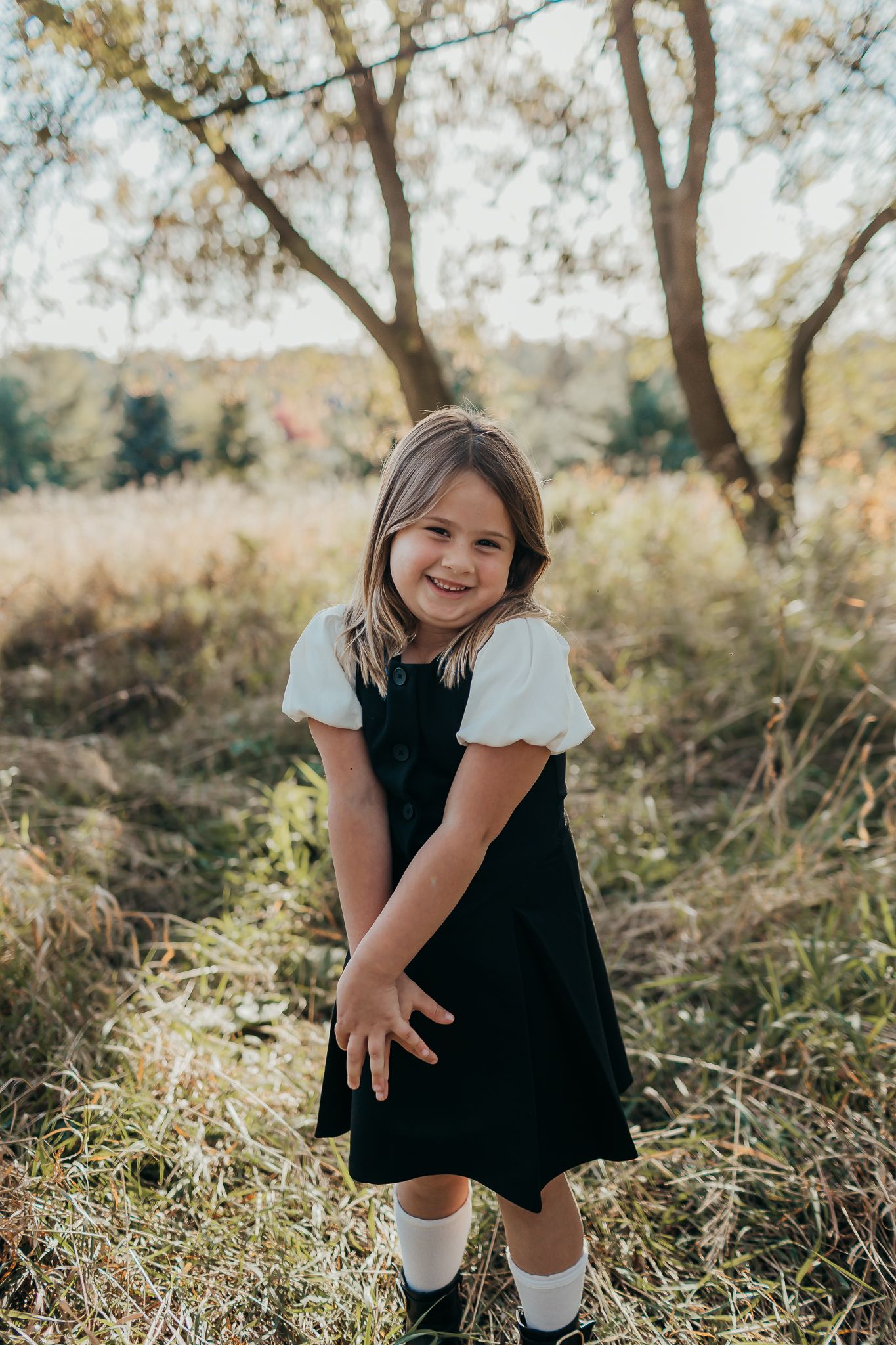 A child in a black dress and white socks poses playfully in an autumn field.
