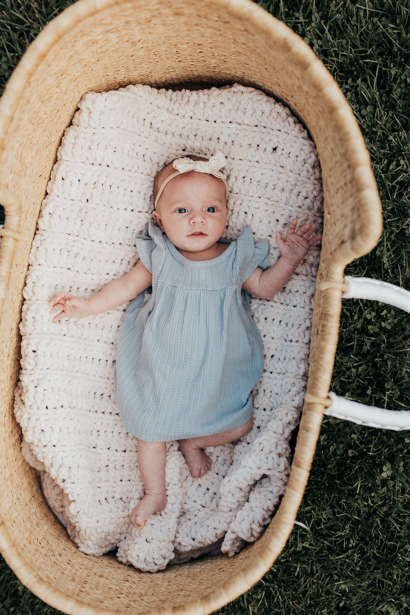 Baby wearing light blue dress lies peacefully in woven basket on white blanket against grass background.