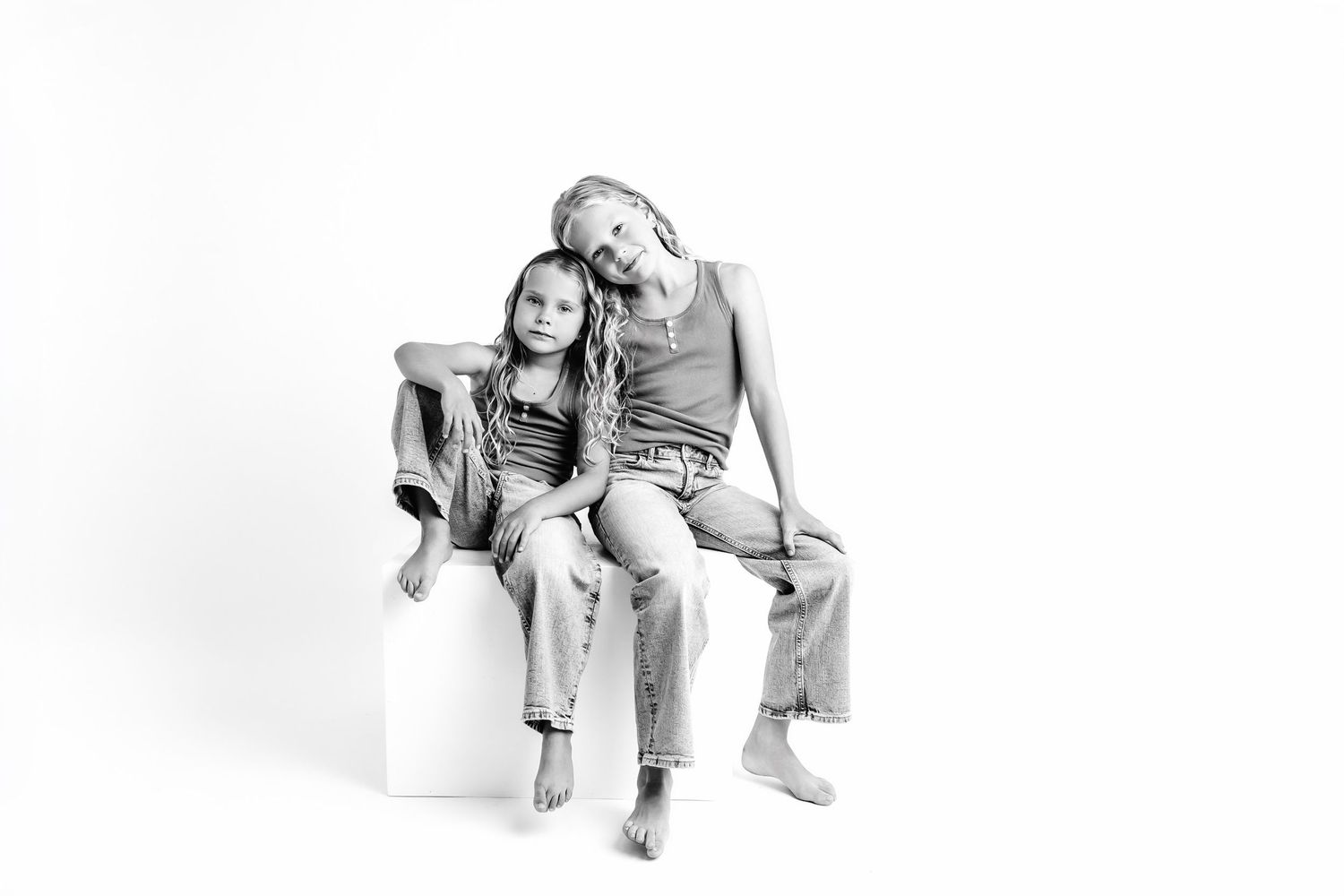 Black and white studio photo of two girls sitting together on a white cube, wearing casual denim outfits.