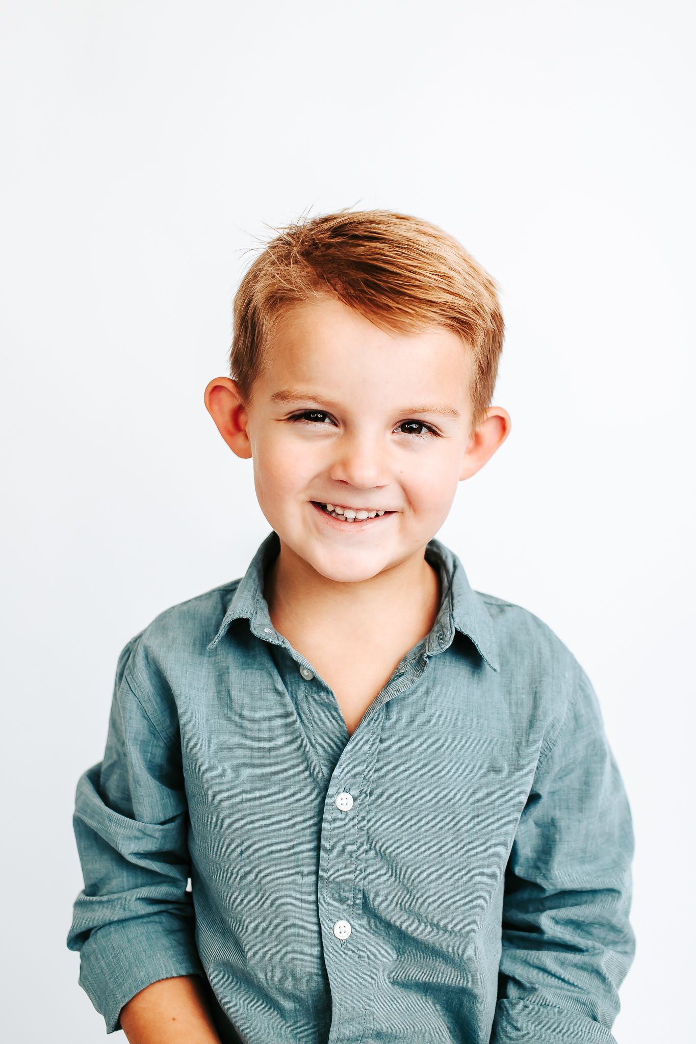 Young child wearing a blue-gray collared shirt smiles warmly in a studio portrait series.
