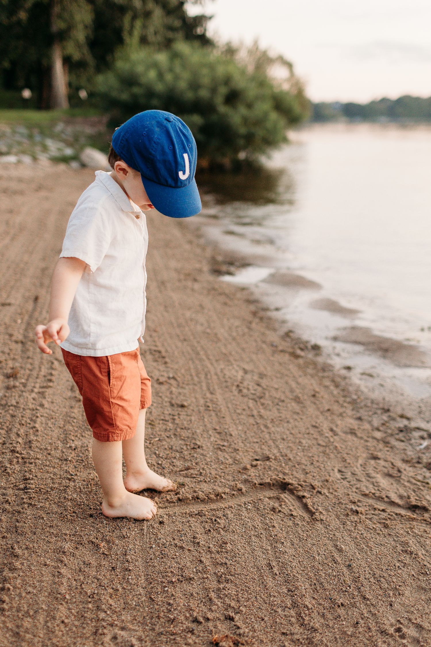 A young child in a blue baseball cap explores a sandy beach shoreline.