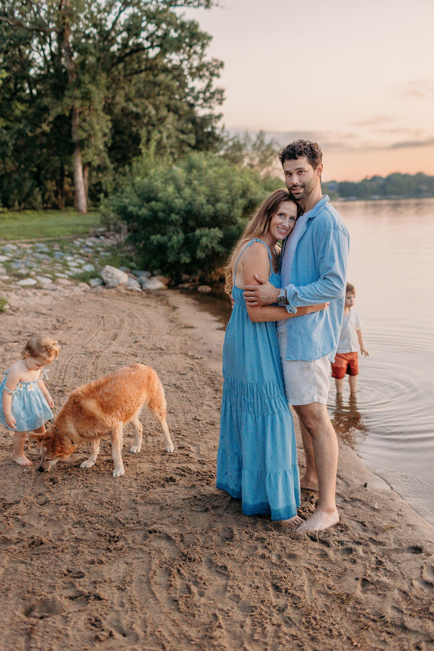 A couple embraces on a beach at sunset while their child plays nearby with a dog.