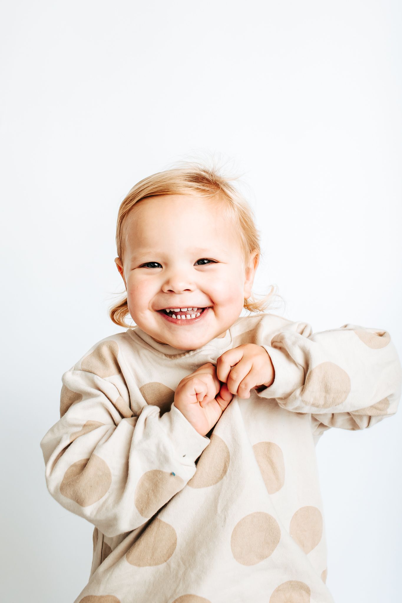 A cheerful toddler in a beige sweater smiles and plays against a white background.