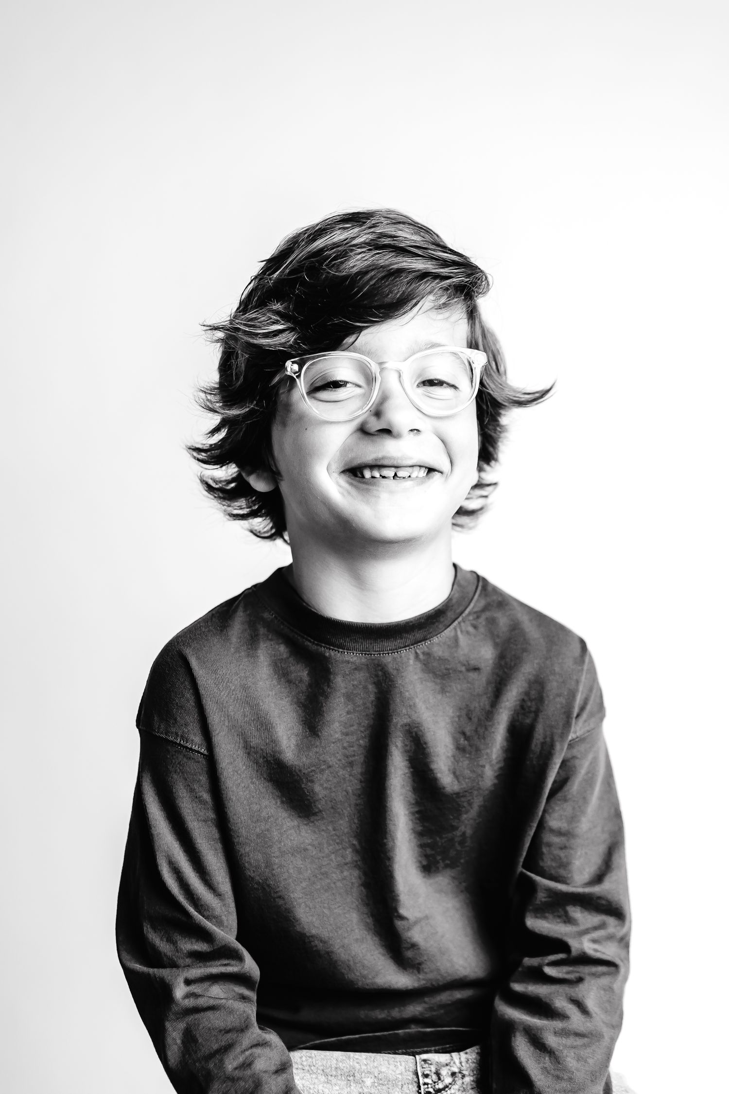 Black and white portrait of a young child wearing glasses and a dark sweater, smiling brightly against a plain background.