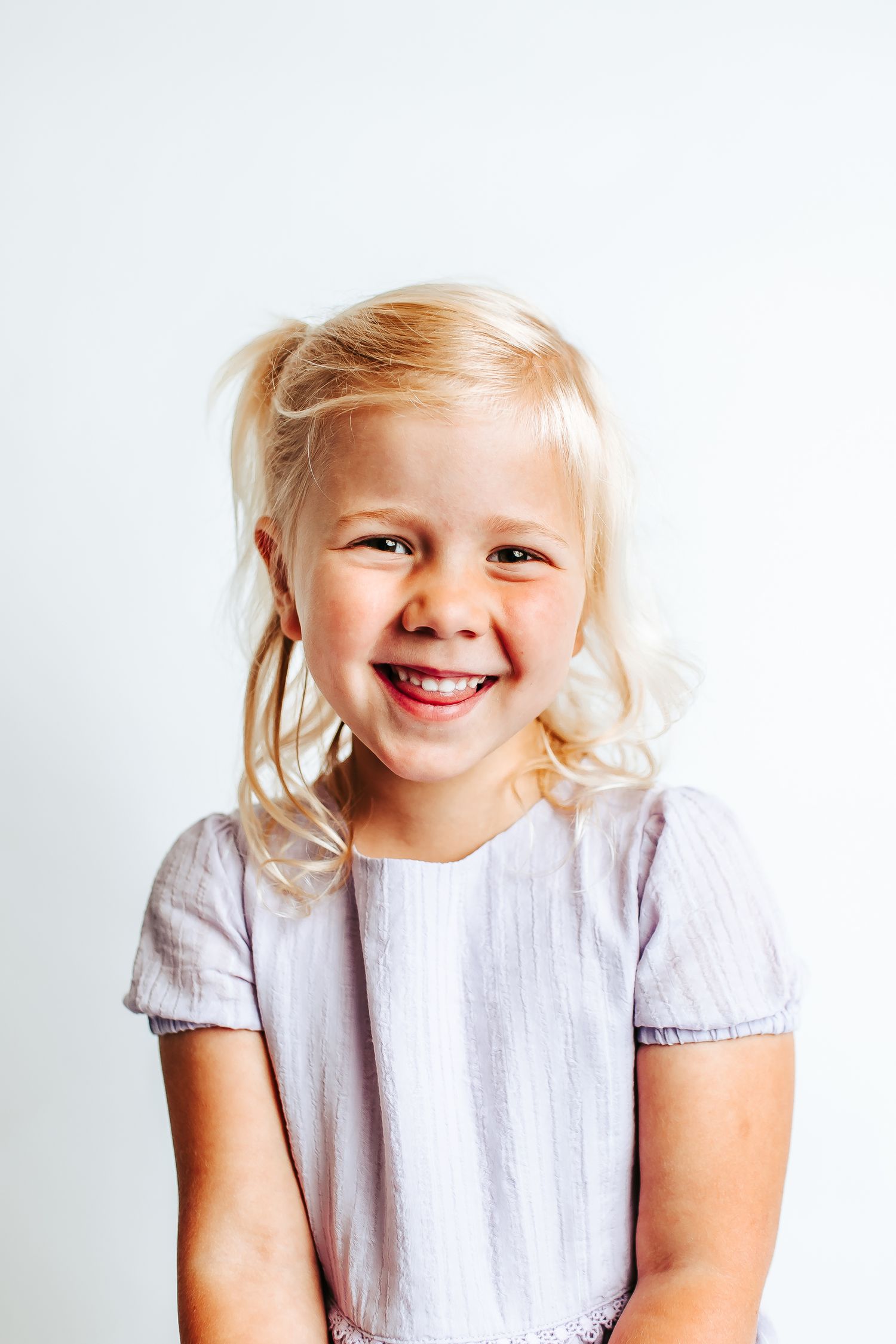 A young child with blonde hair wearing a white dress smiles brightly against a light background.