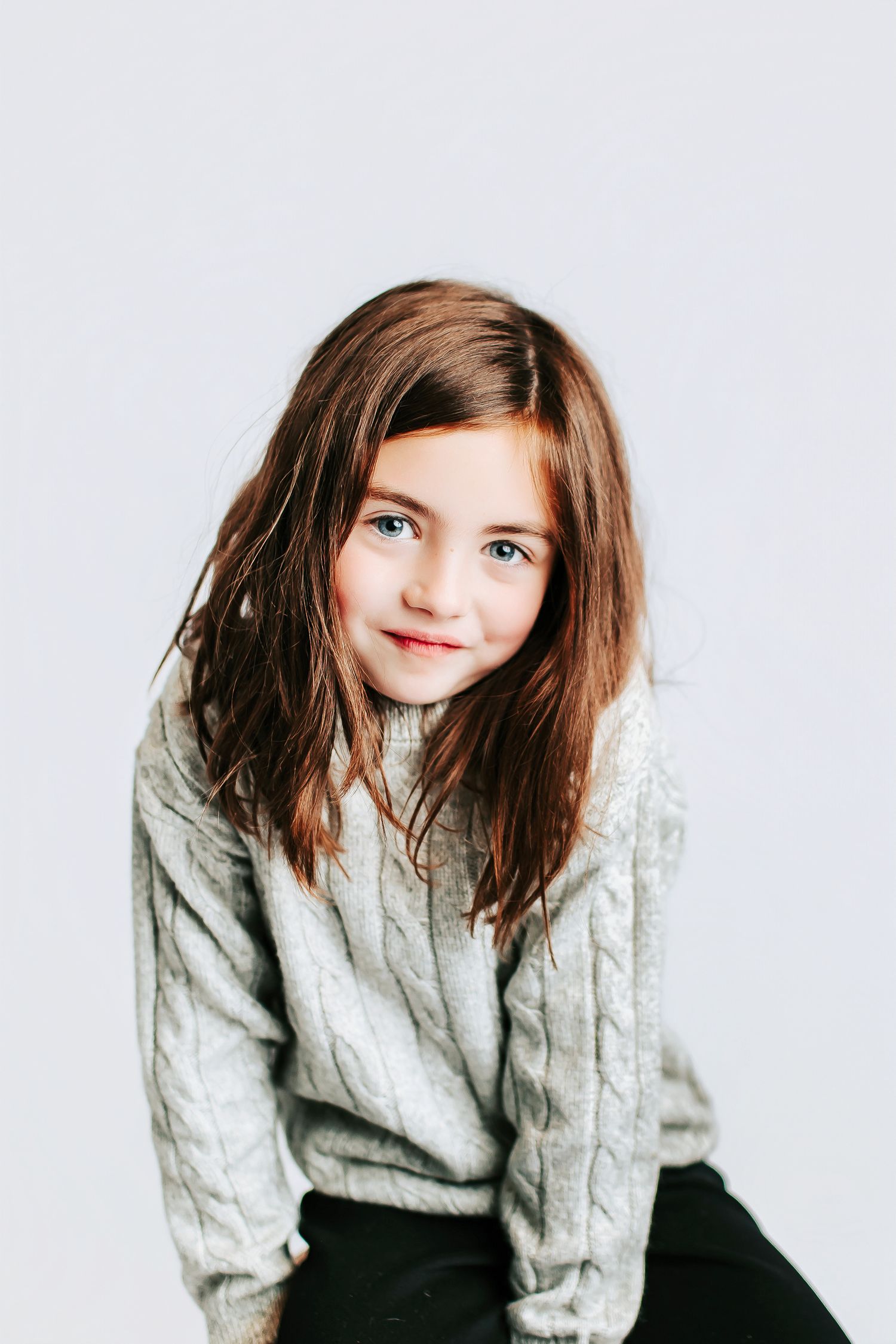 Young girl in gray sweater poses against white background with natural expression.