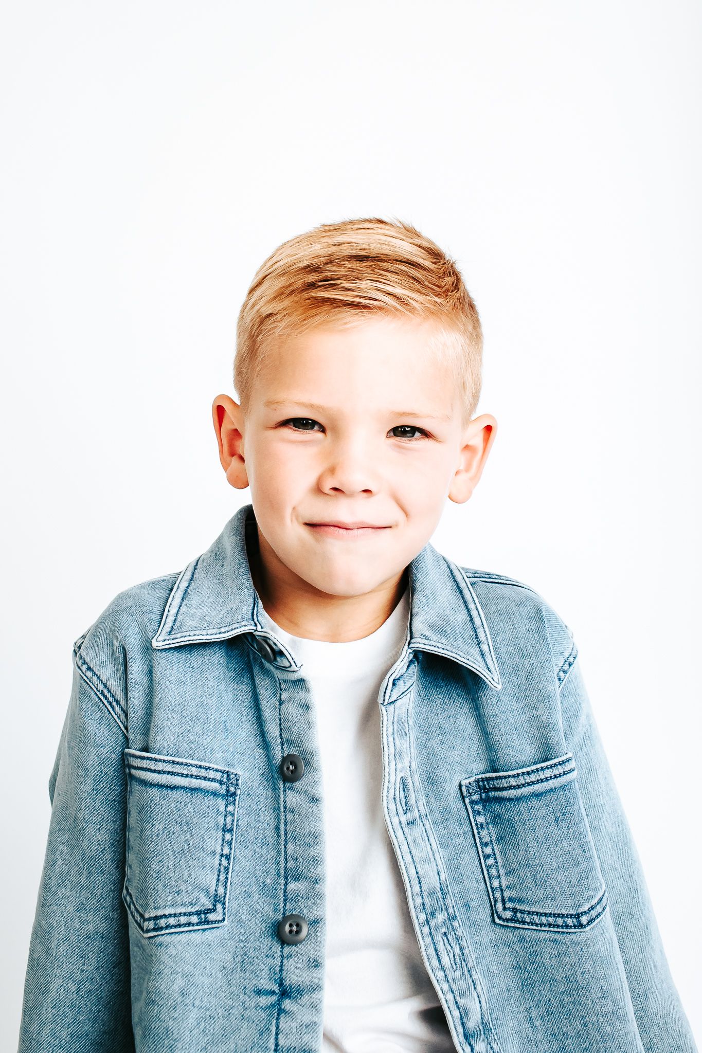 Young child wearing denim jacket and white shirt smiles against white background.