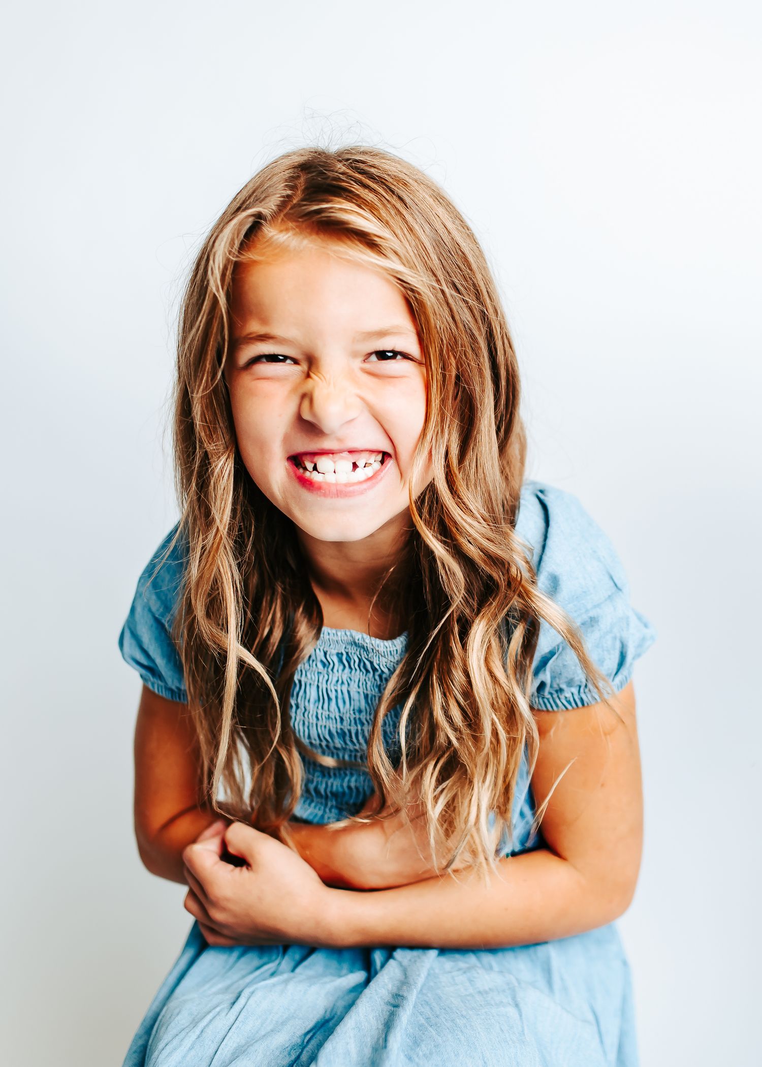 Young child wearing blue dress smiles joyfully with wavy blonde hair against white background.