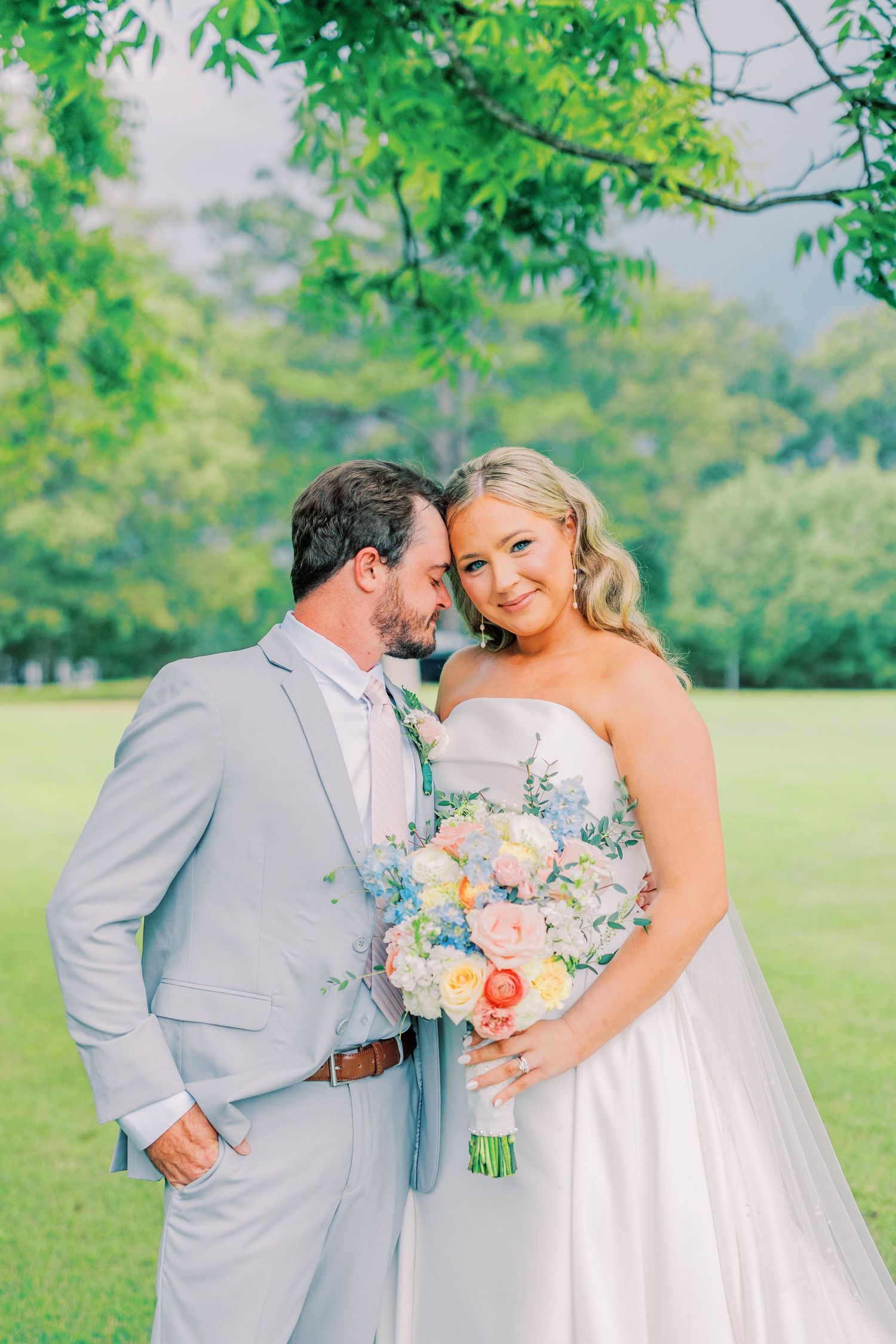 Bride and groom share a romantic moment outdoors, she holds a pastel bouquet, he wears a light gray suit.