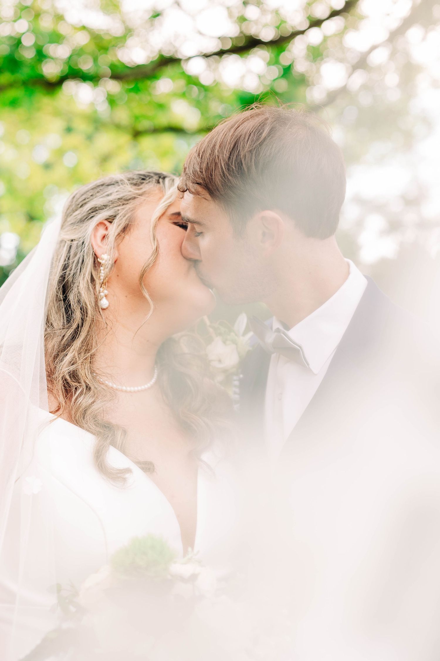 A romantic wedding couple shares an intimate kiss surrounded by soft white light and greenery.