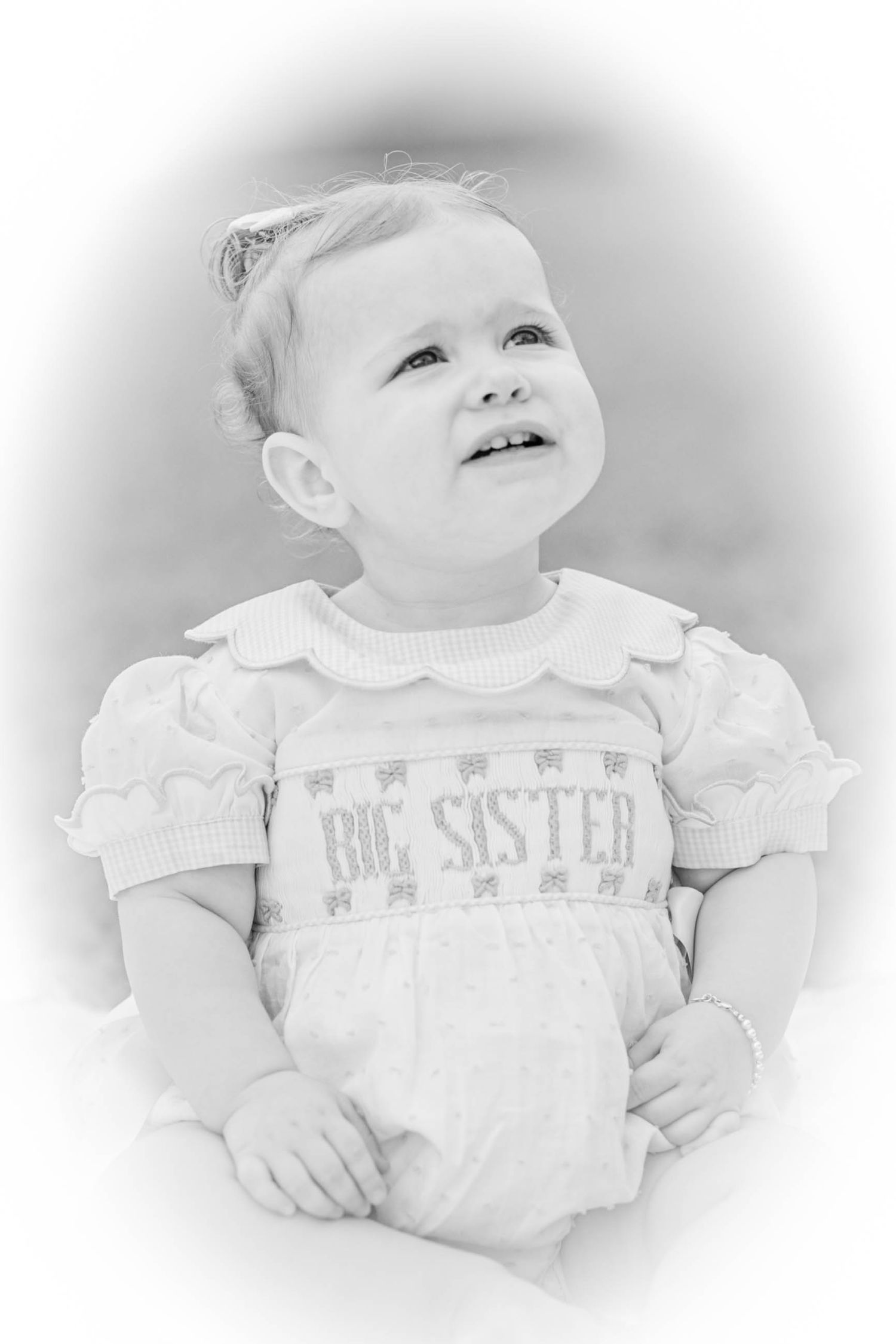 Black and white portrait of a toddler girl wearing a smocked Big Sister outfit, looking up with a sweet smile.