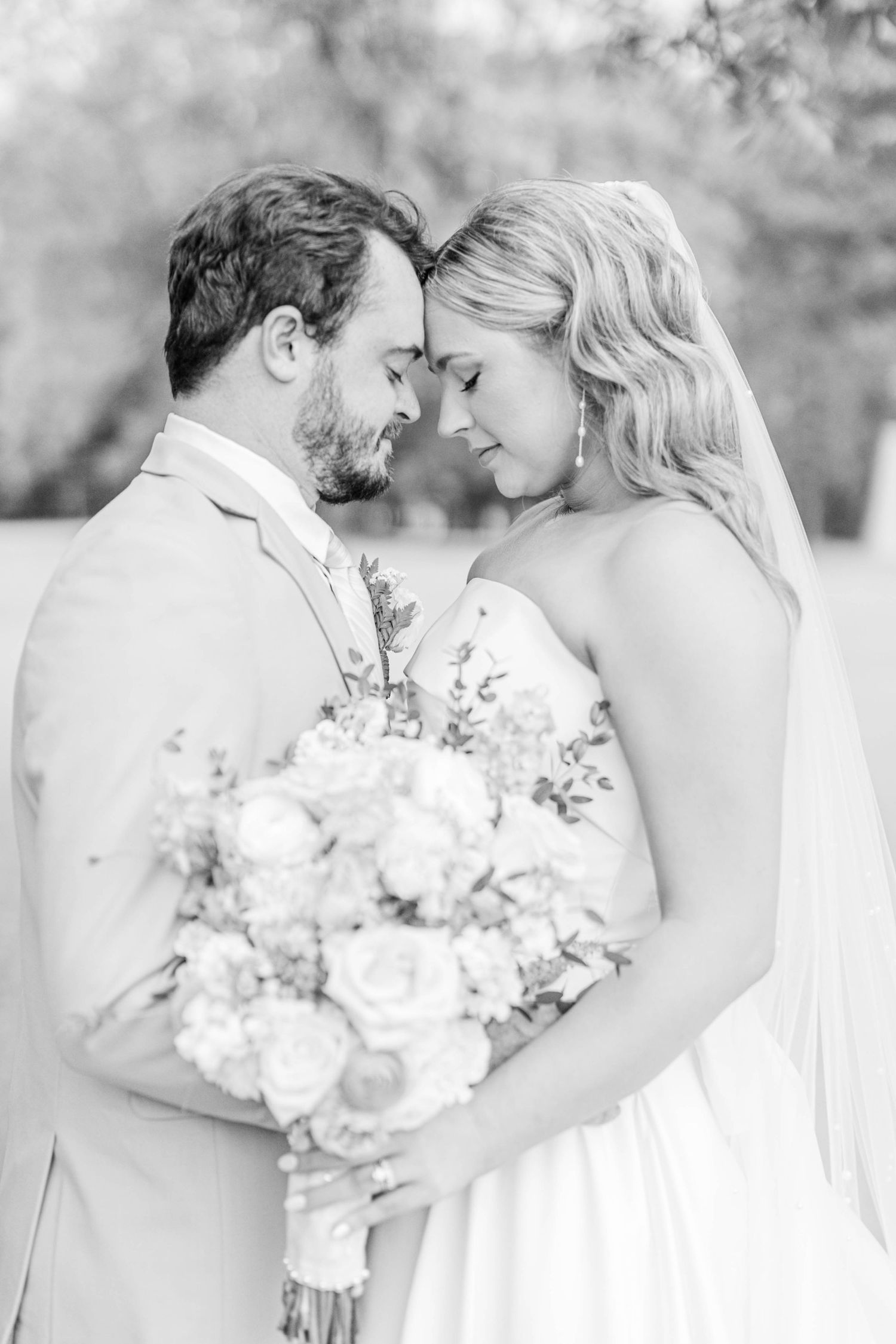 Black and white photo of a bride and groom touching foreheads, bride holding a floral bouquet.