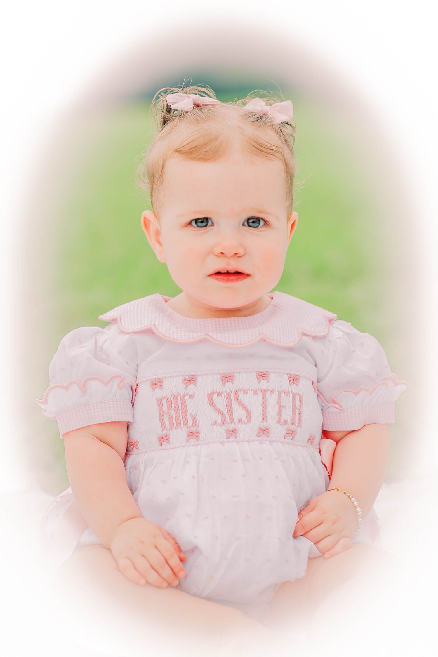 Toddler girl in pink Big Sister smocked dress with bow, posed for portrait photo outdoors.