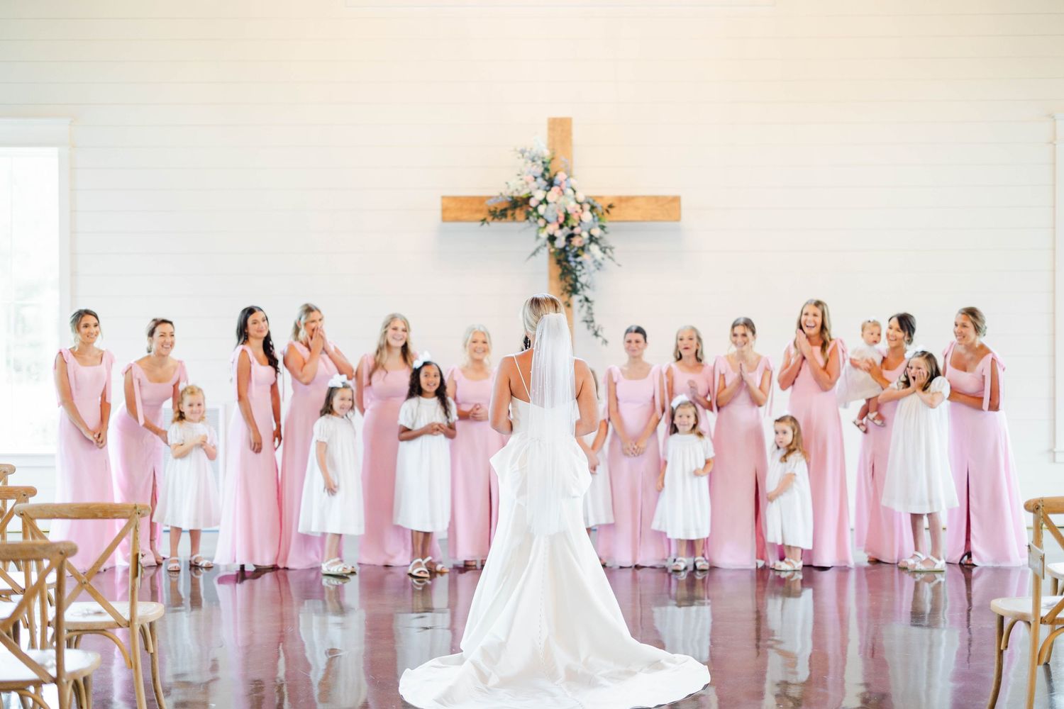 Wedding party wearing pink bridesmaid dresses pose in front of wooden cross with floral arrangement.