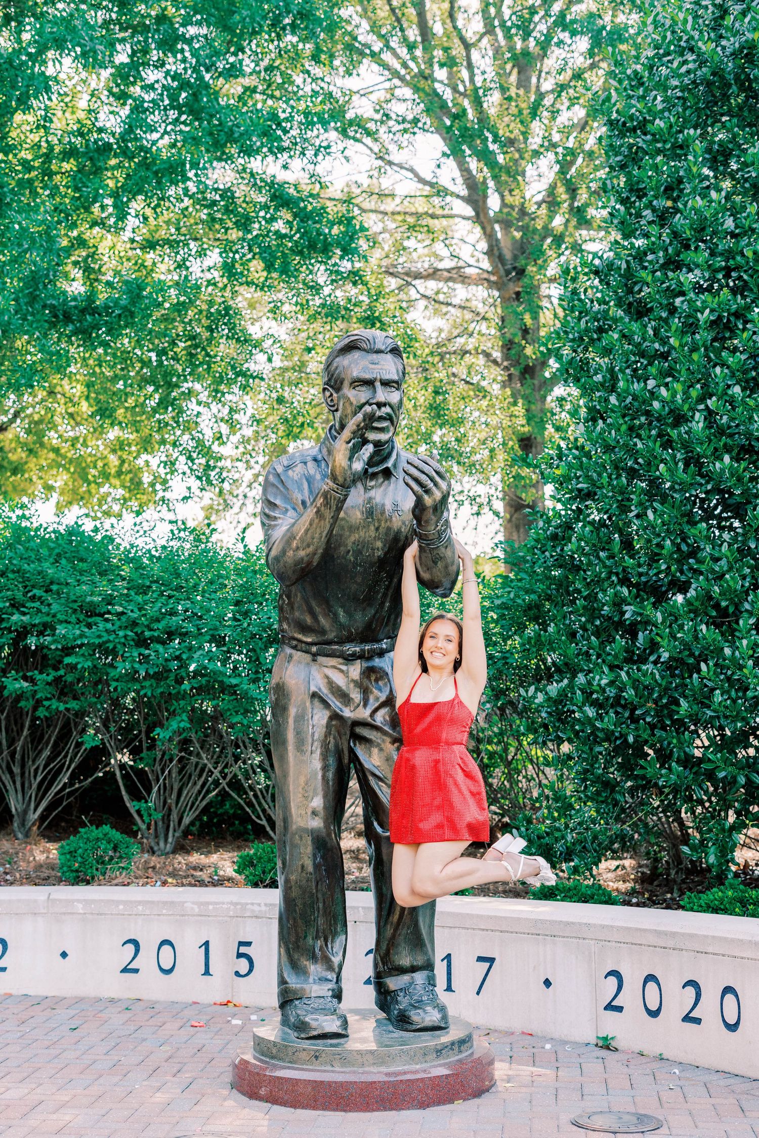 Woman in red dress poses beside a bronze statue in a garden with lush green trees and commemorative year markers.