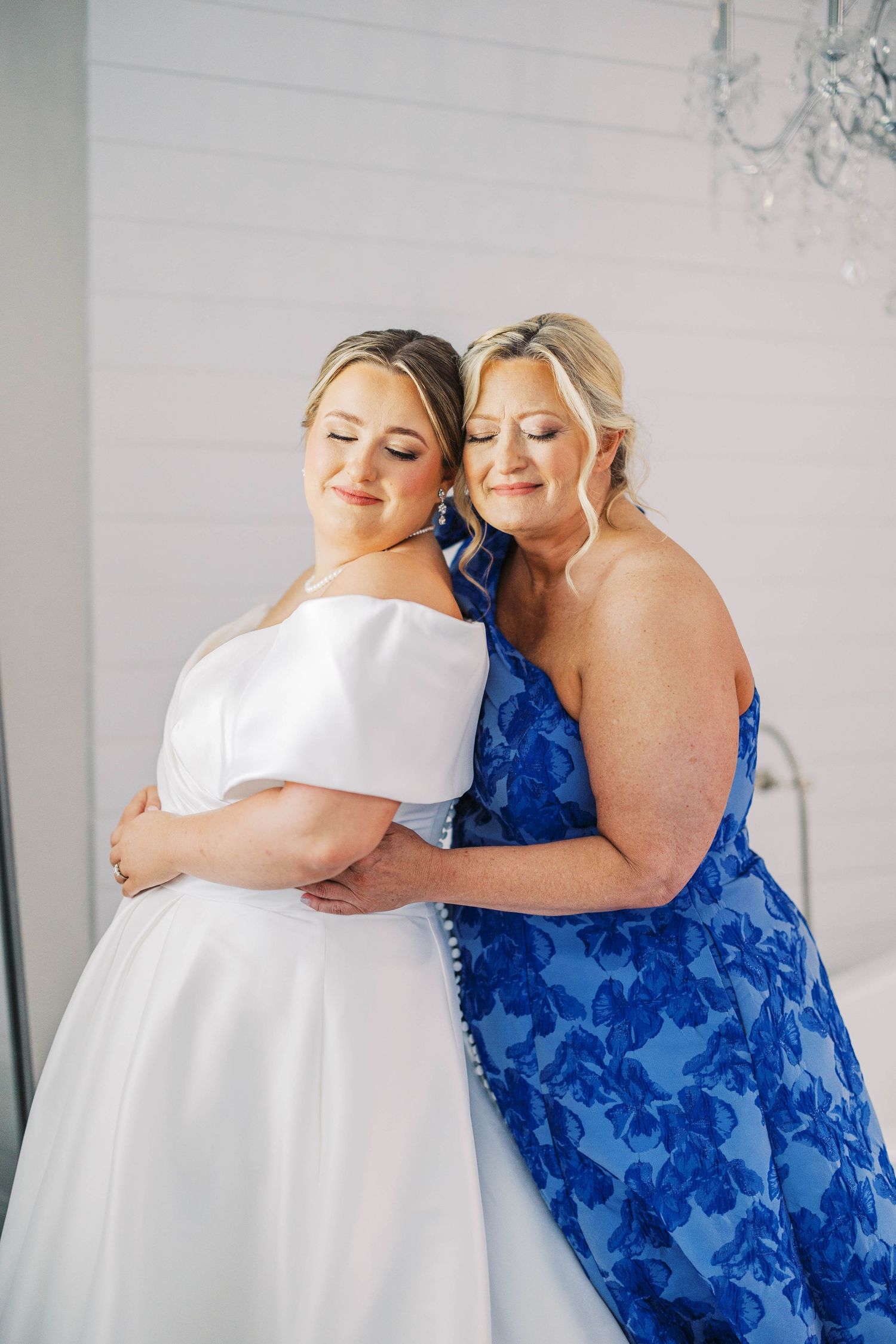 Emotional embrace between woman in white dress and another in blue floral gown.