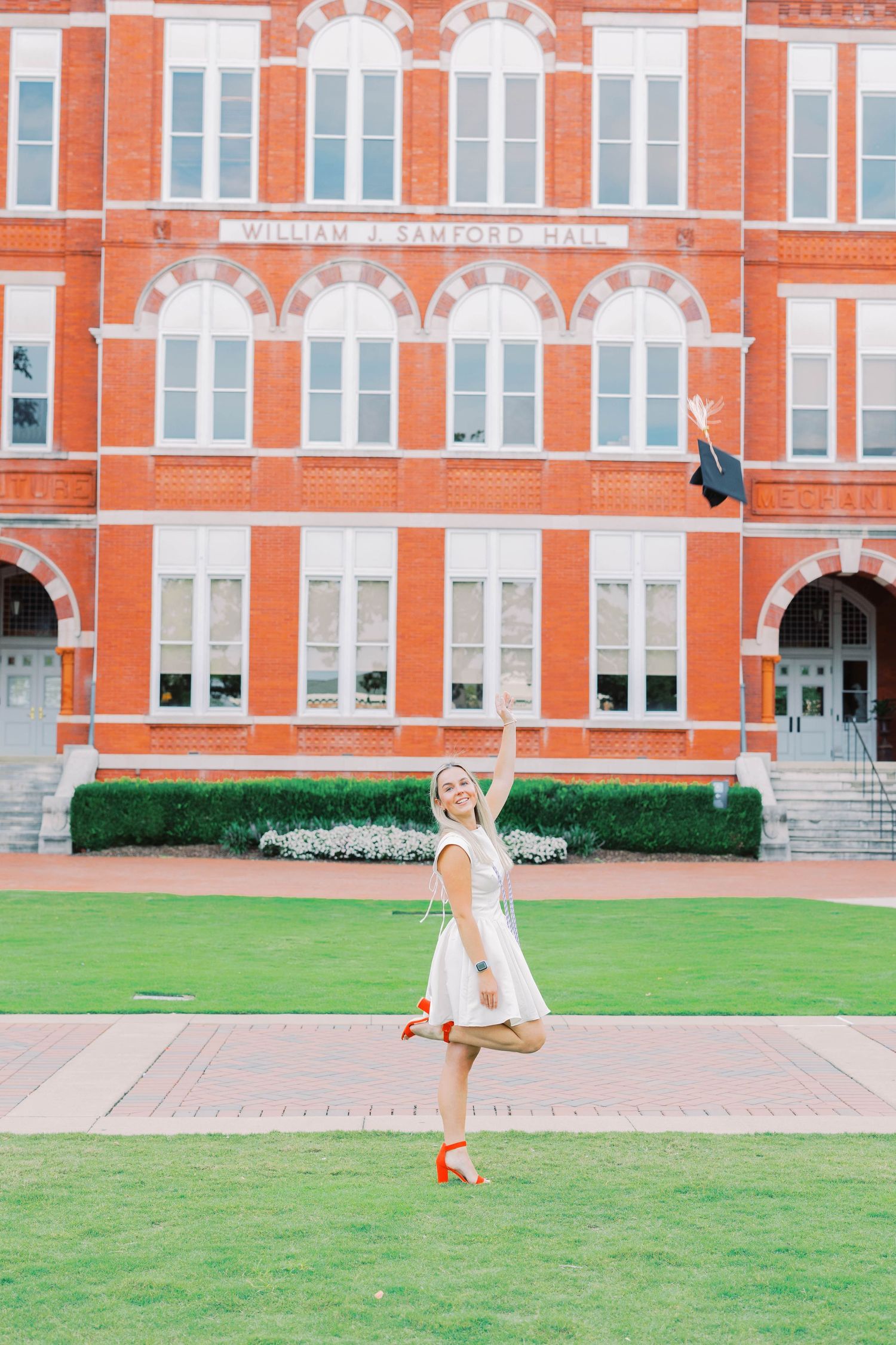 A person in white poses on a lawn in front of a grand red brick building.