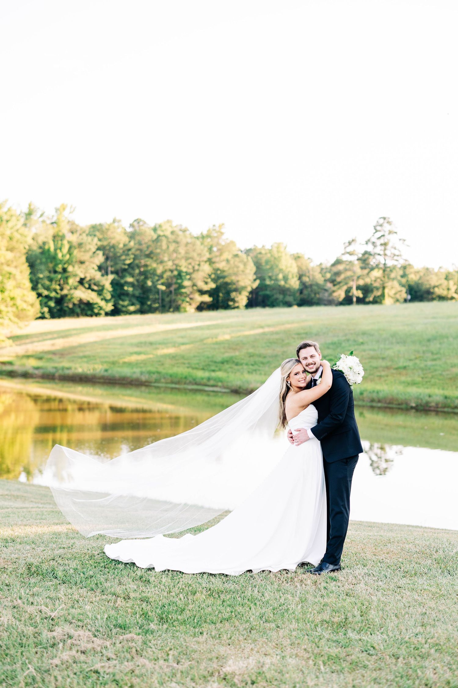 Romantic wedding portrait of couple embracing by a lake with flowing white veil.