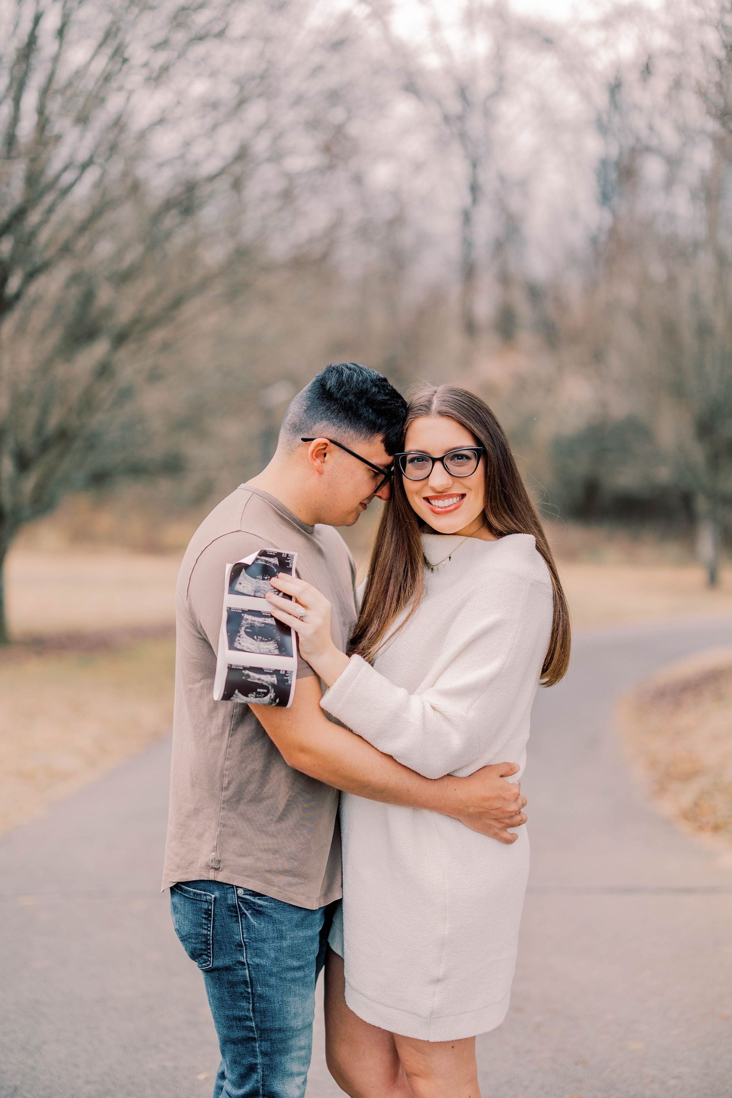 Expecting couple embracing while holding ultrasound photos on outdoor path.