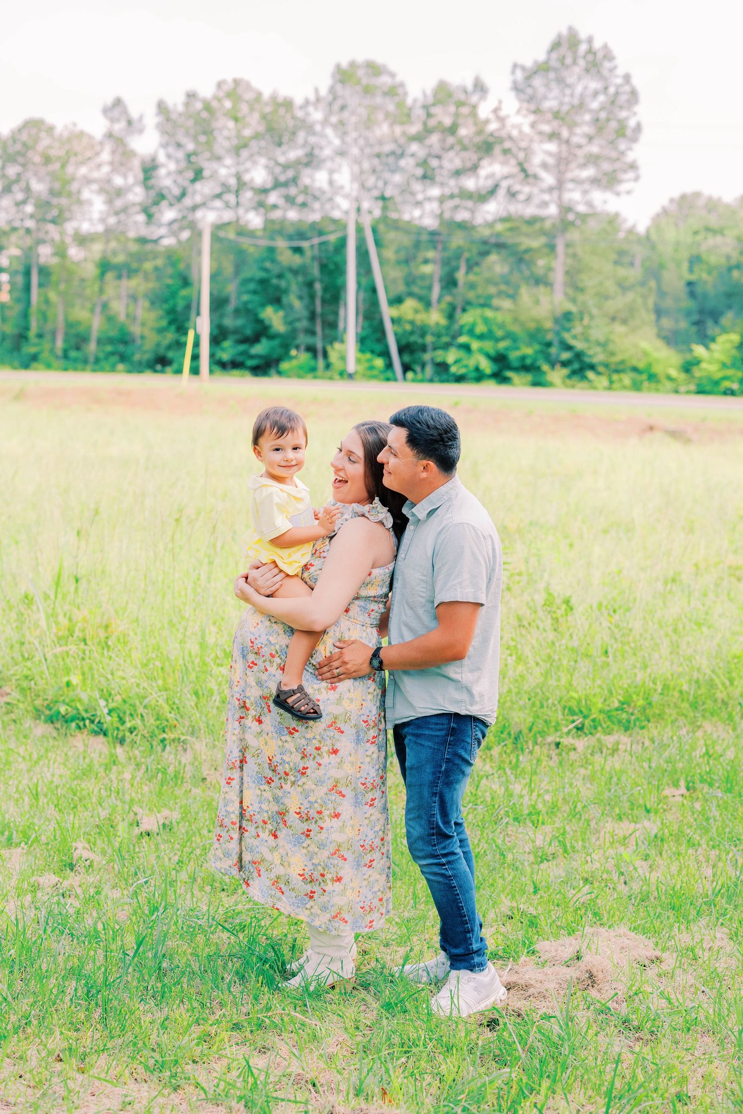 A happy family of three poses in a green field, with parents holding their toddler outdoors near tall trees.