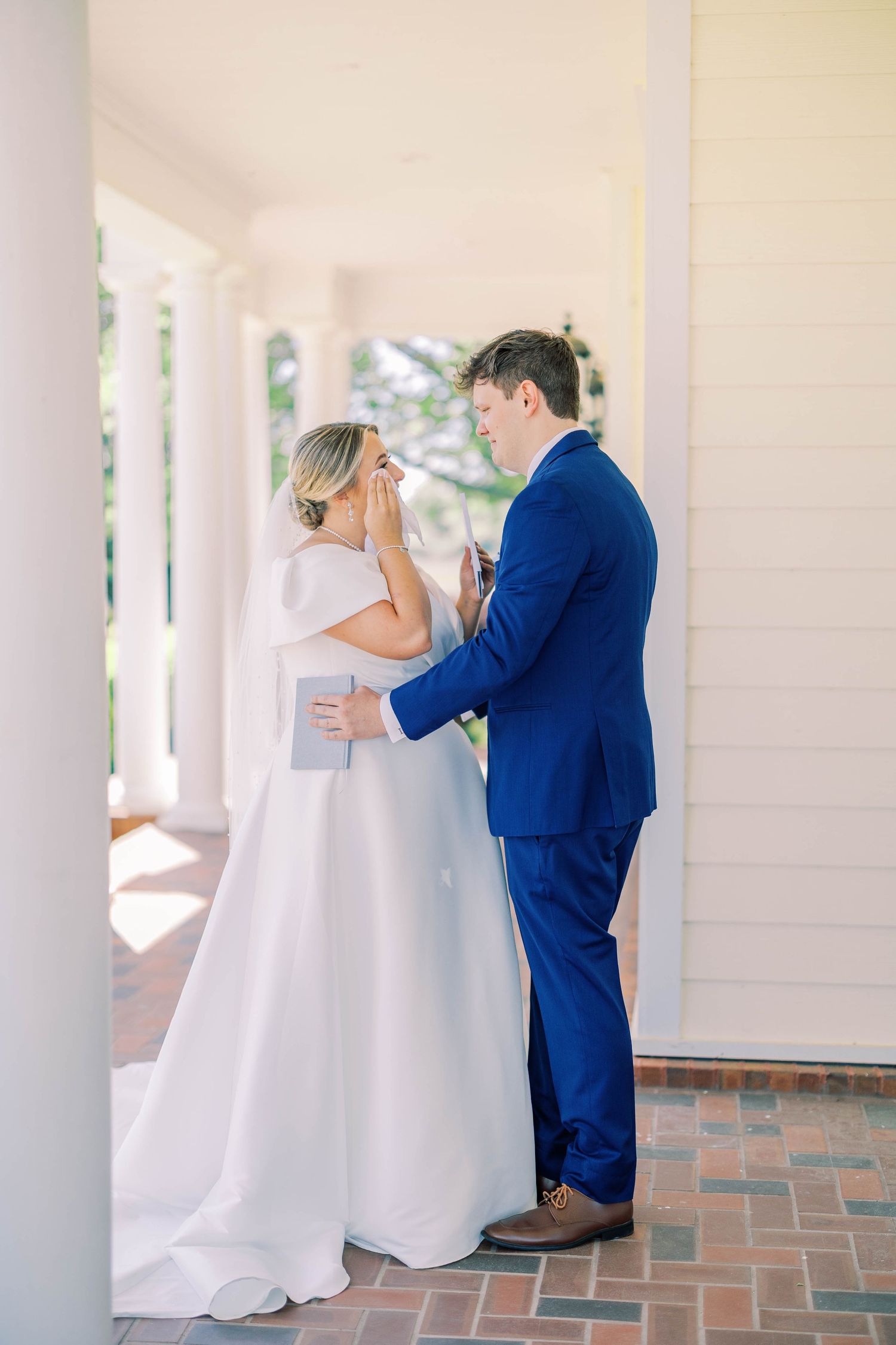 A tender first look moment between a couple in a white dress and blue suit under a columned porch.