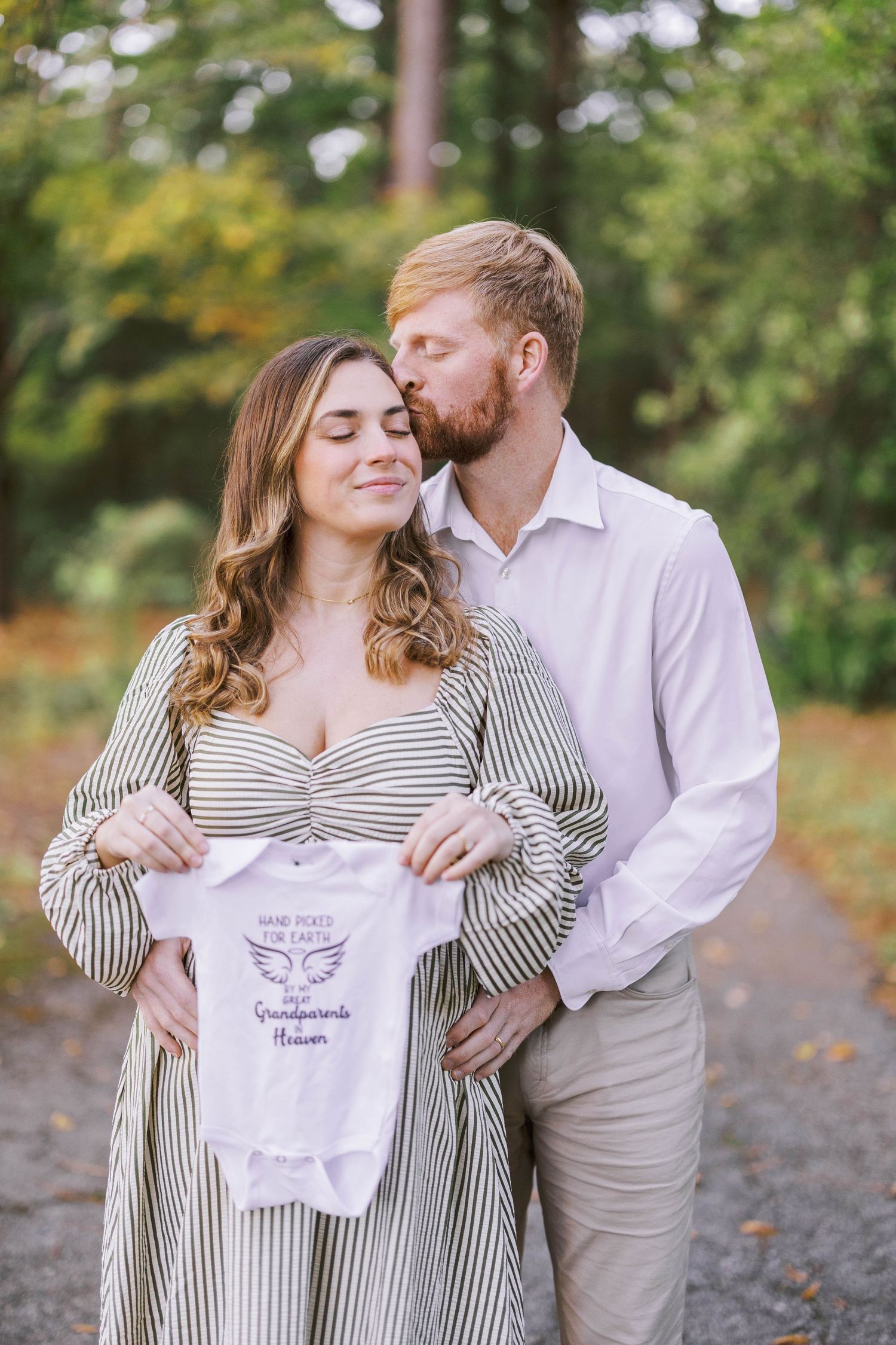 A couple displays a baby onesie while sharing an intimate moment outdoors.