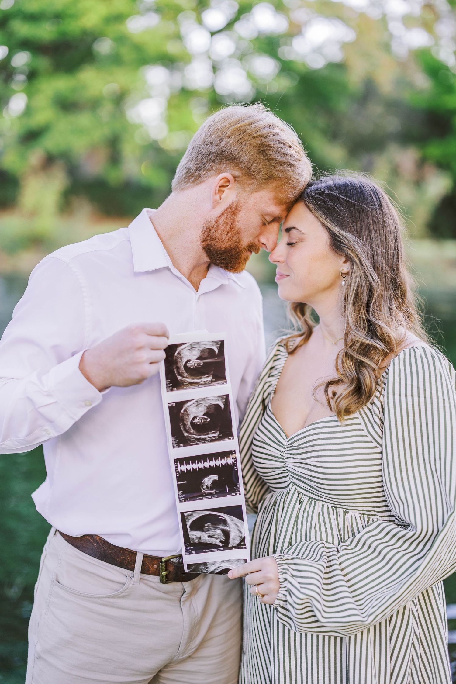 A couple shares an intimate moment while holding ultrasound photos in a park setting.