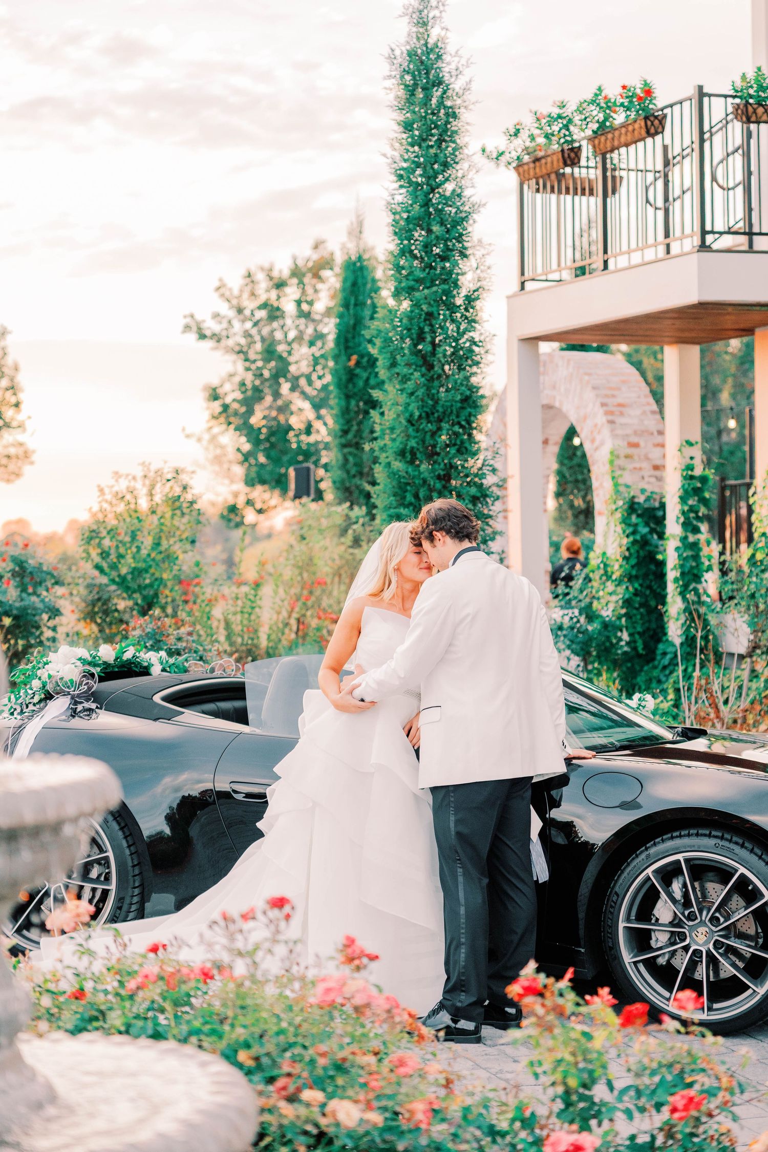 A romantic wedding moment captured at sunset beside a luxury sports car with cypress trees and blooming flowers in the background.