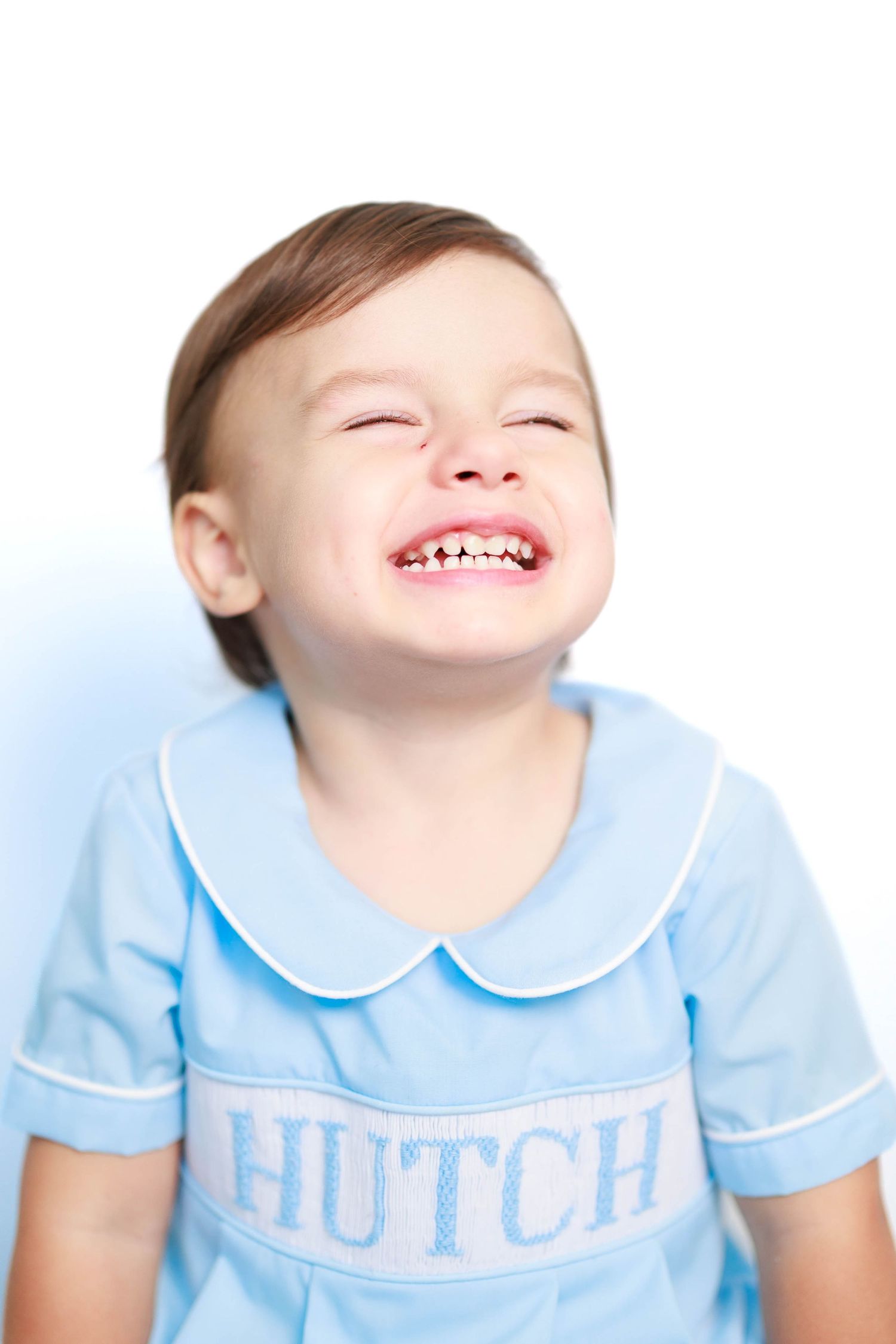 Laughing toddler boy wearing a blue smocked outfit embroidered with the name Hutch against a white background.