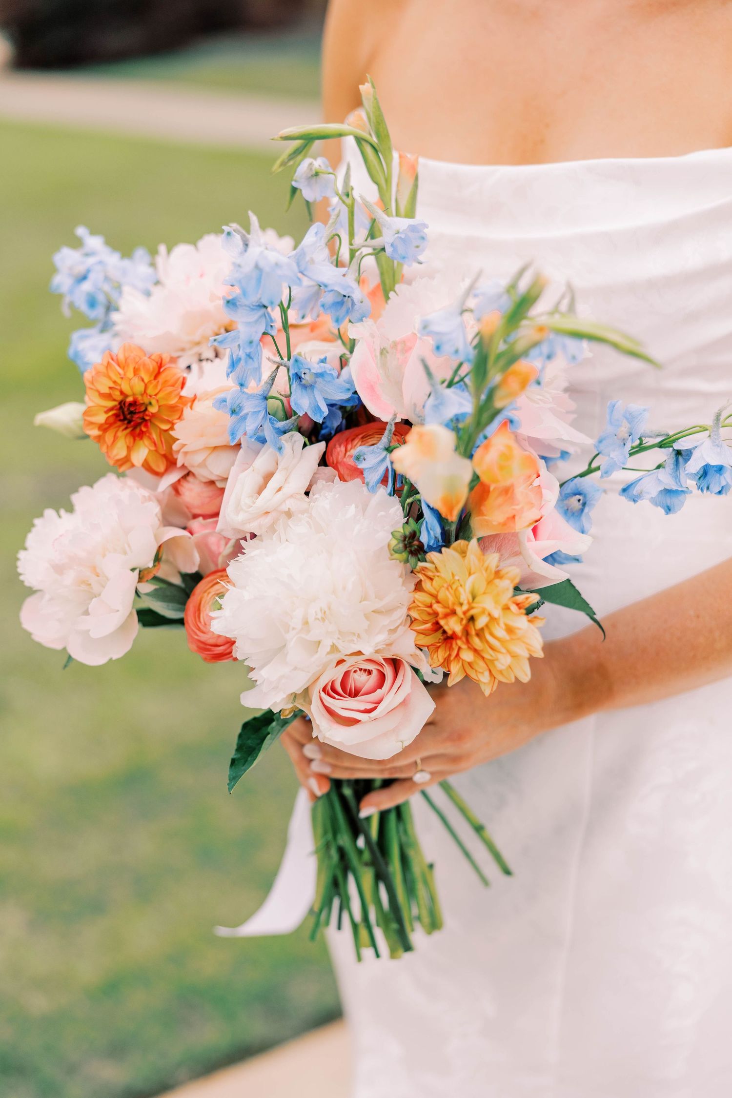 A romantic wedding bouquet featuring pink roses, blue delphiniums, orange dahlias, and white peonies against a white dress.
