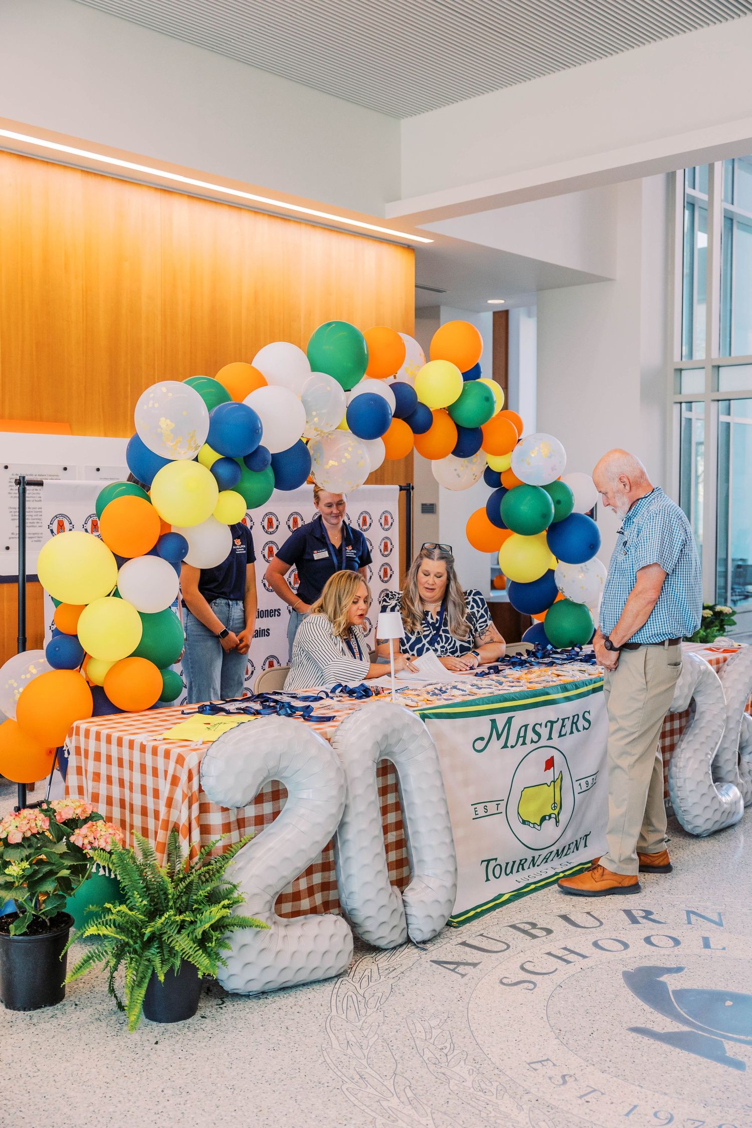 Masters Tournament themed event with balloon arch in green, blue, orange and yellow at Auburn School table with 2024 balloon numbers.