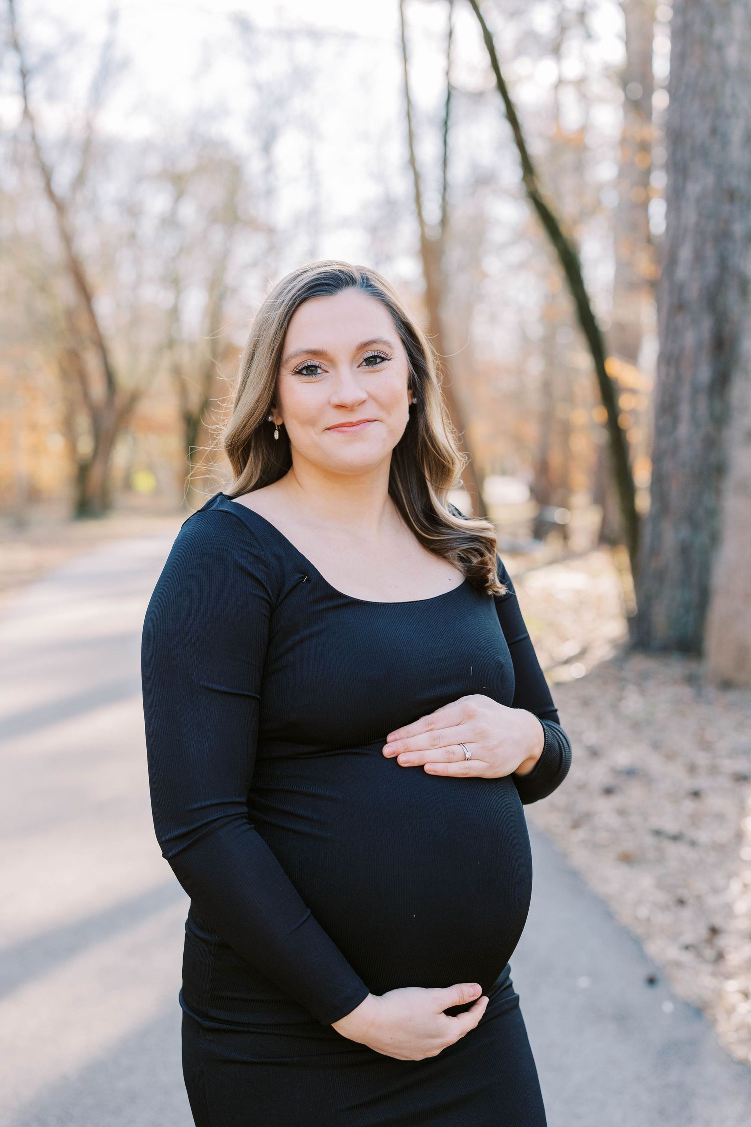 Woman in black long sleeve dress cradling baby bump outdoors.