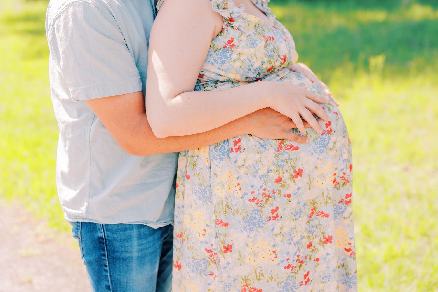 Couple embracing outdoors, pregnant woman in floral dress with partner in blue shirt and jeans on sunny day.