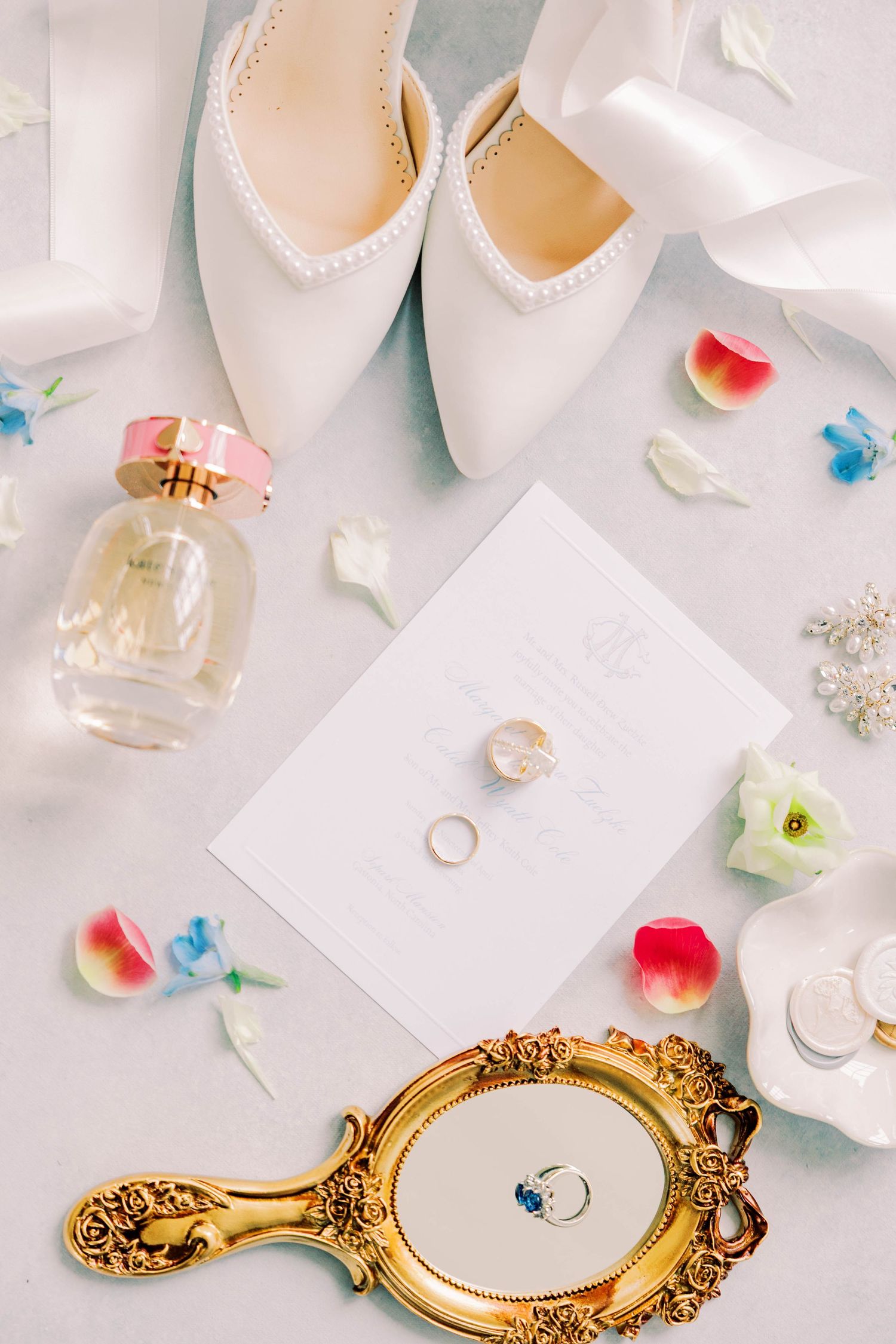 Elegant wedding flat lay with white heels, gold rings, perfume bottle, ornate mirror, and colorful flower petals on light background.