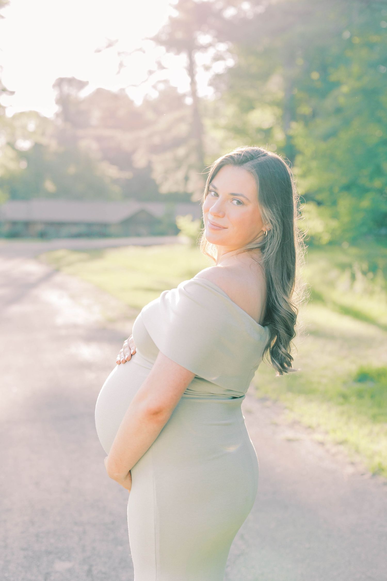 Pregnant woman in sage off-shoulder gown cradling baby bump outdoors in golden sunlight.