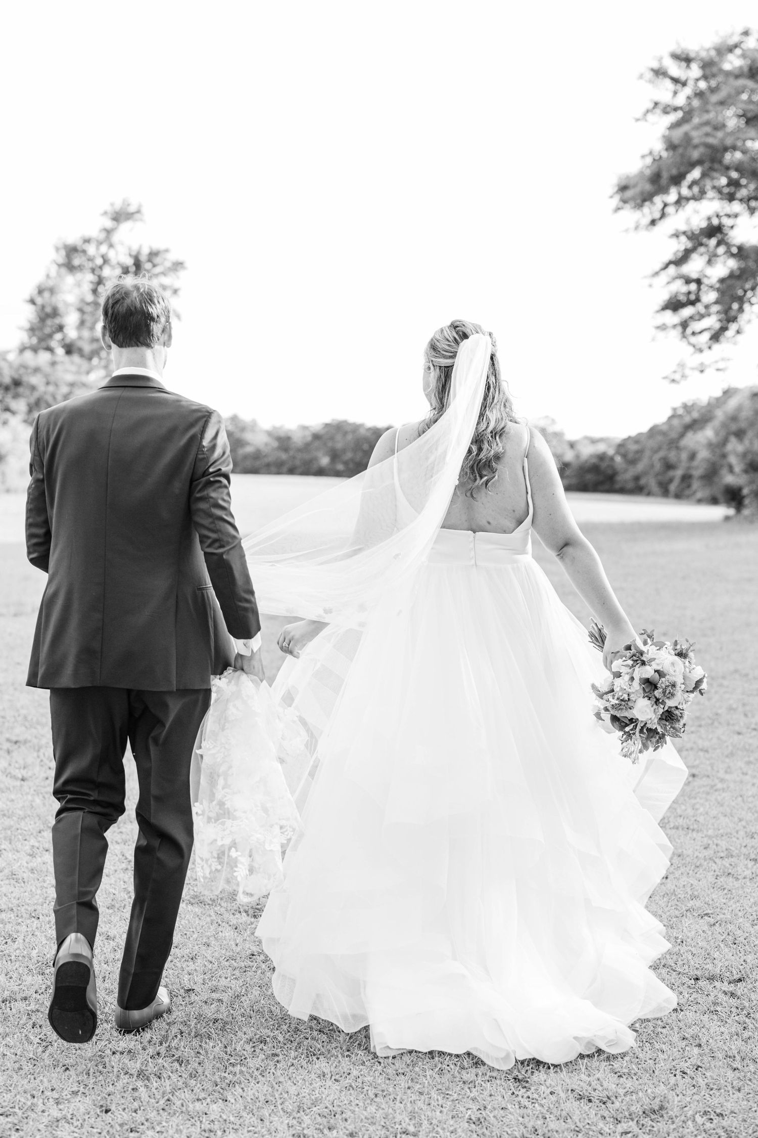 Black and white photo of couple walking together across grassy field in wedding attire.